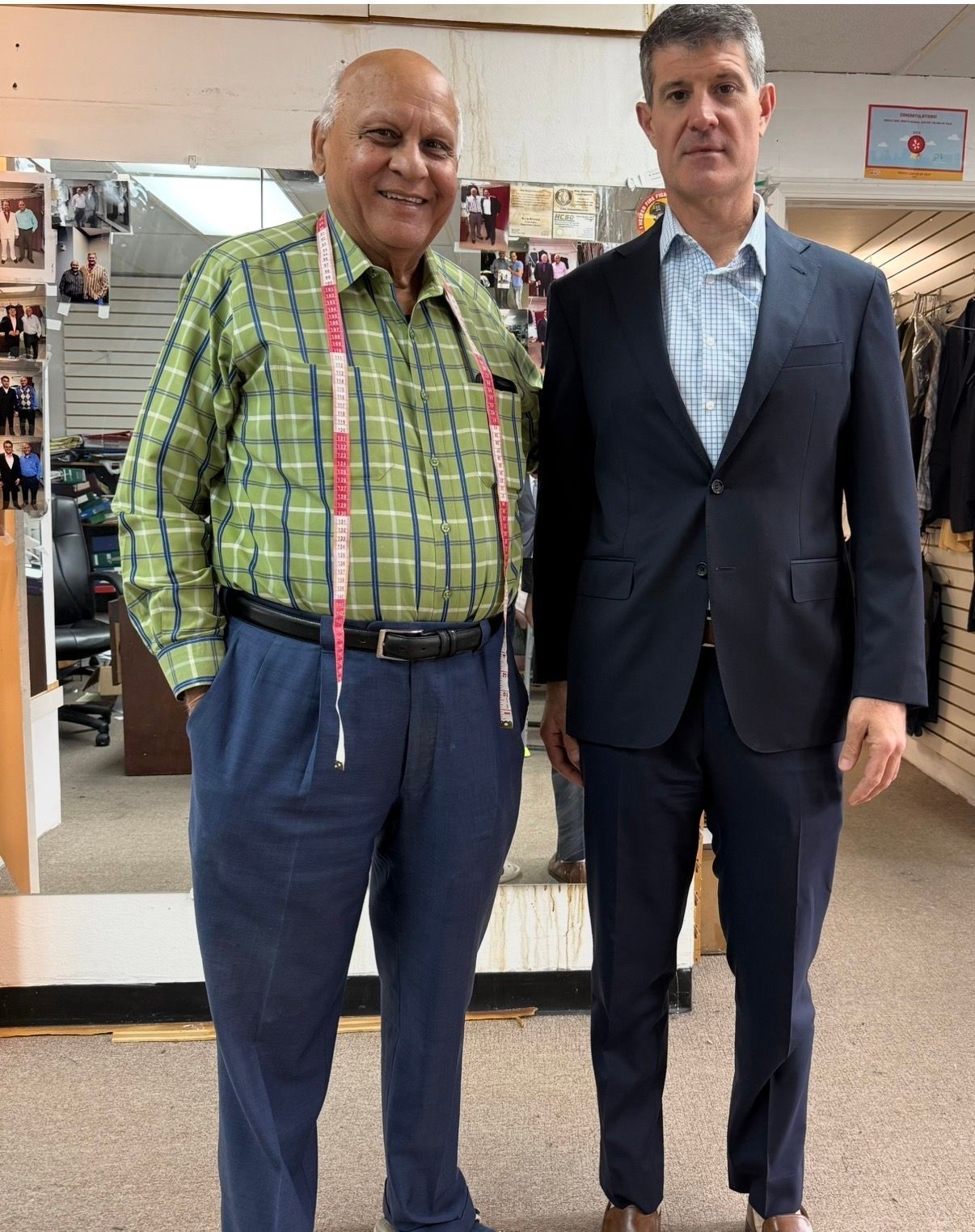 Two men standing together in a clothing store, one in a green plaid shirt and one in a dark suit.