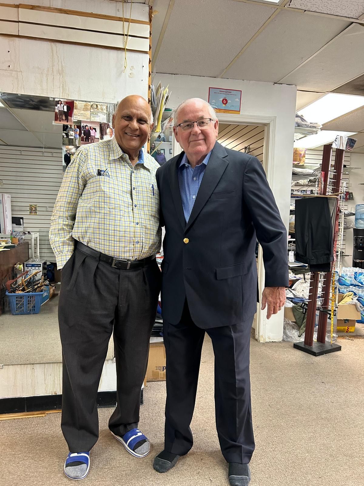 Two men standing together indoors, smiling in a shop with shelves and displays behind them.