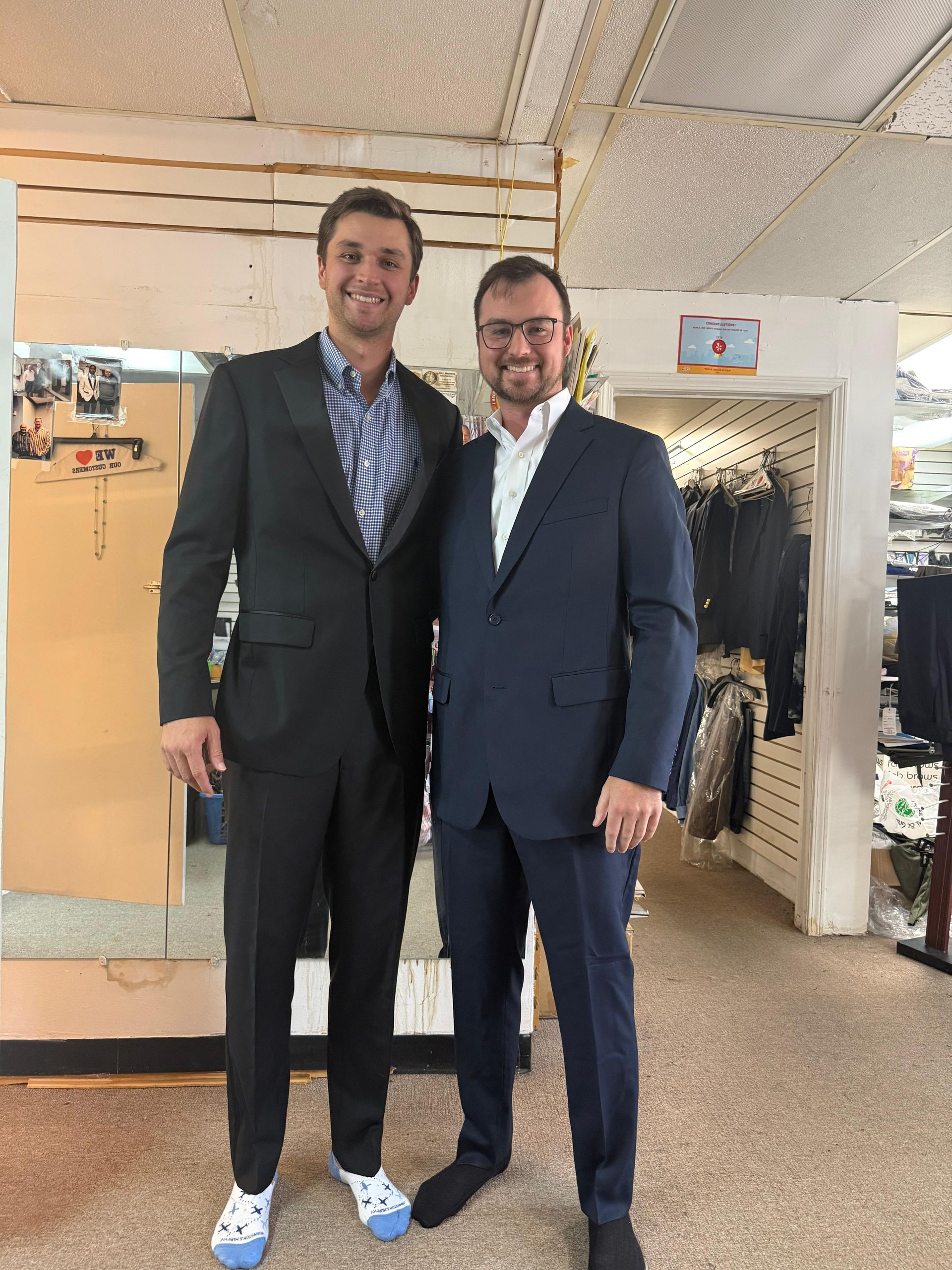 Two men in suits standing together in a clothing store, smiling at the camera.