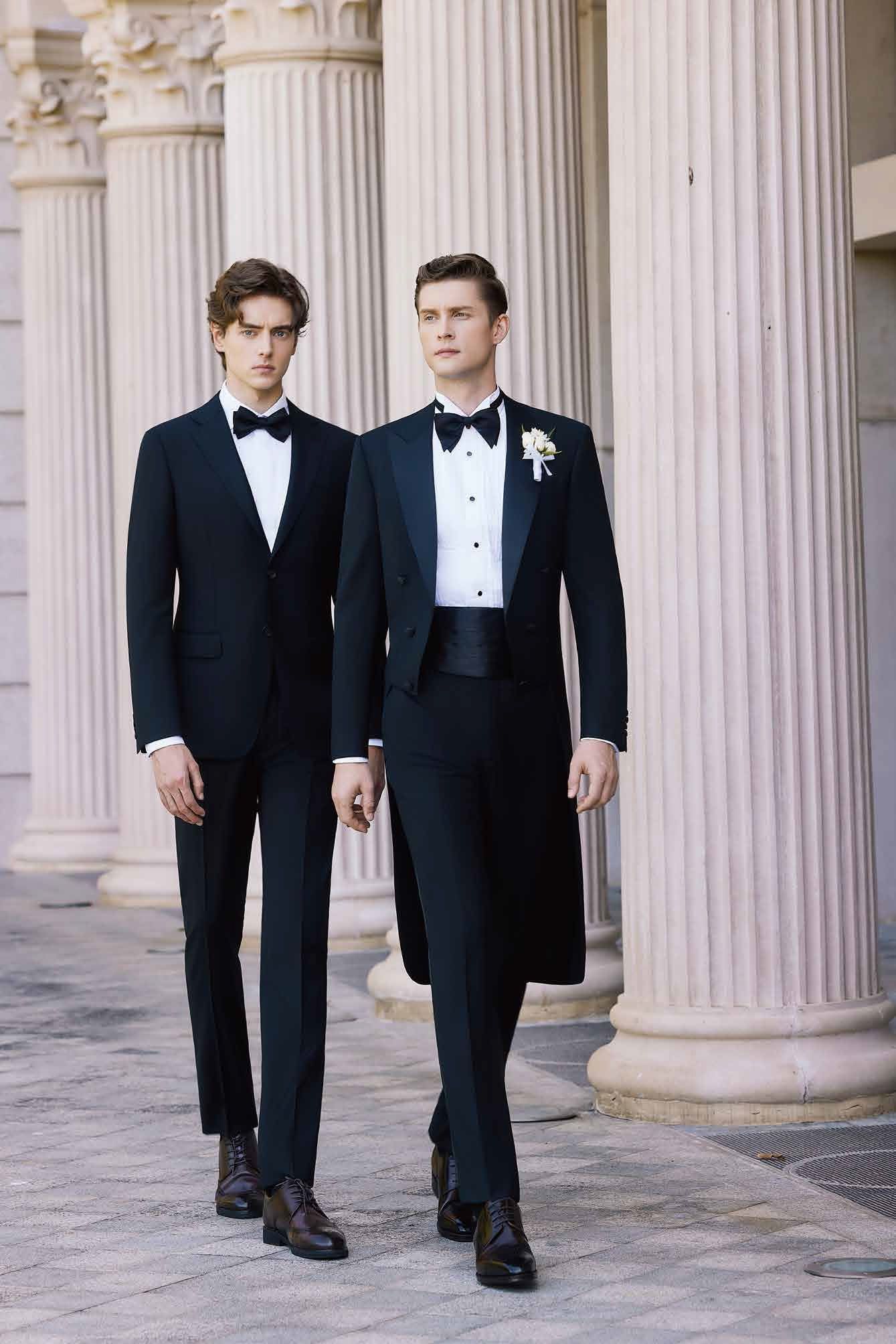 Two men in tuxedos and bow ties are walking in front of columns.