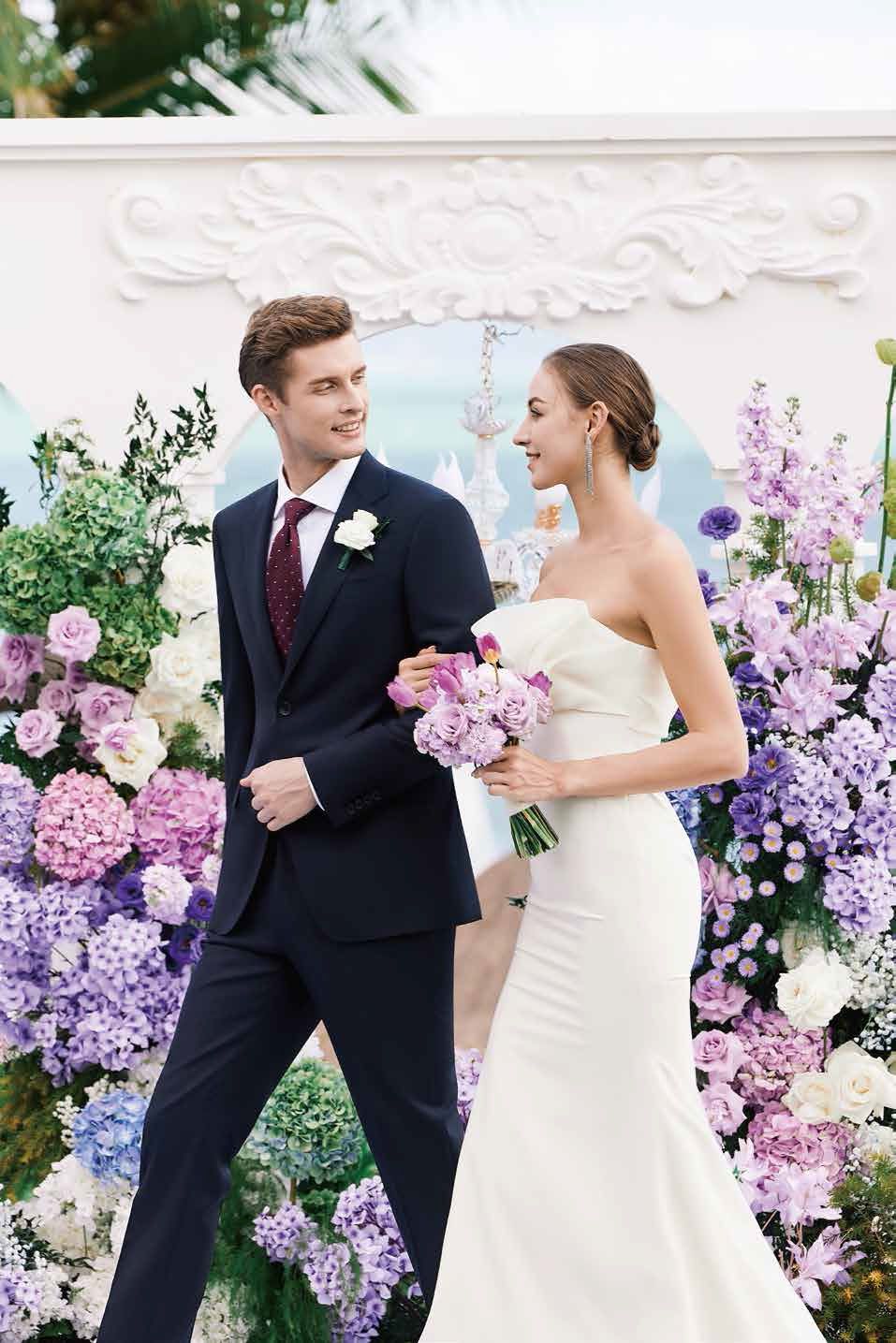 A bride and groom are walking in front of a wall of purple flowers.