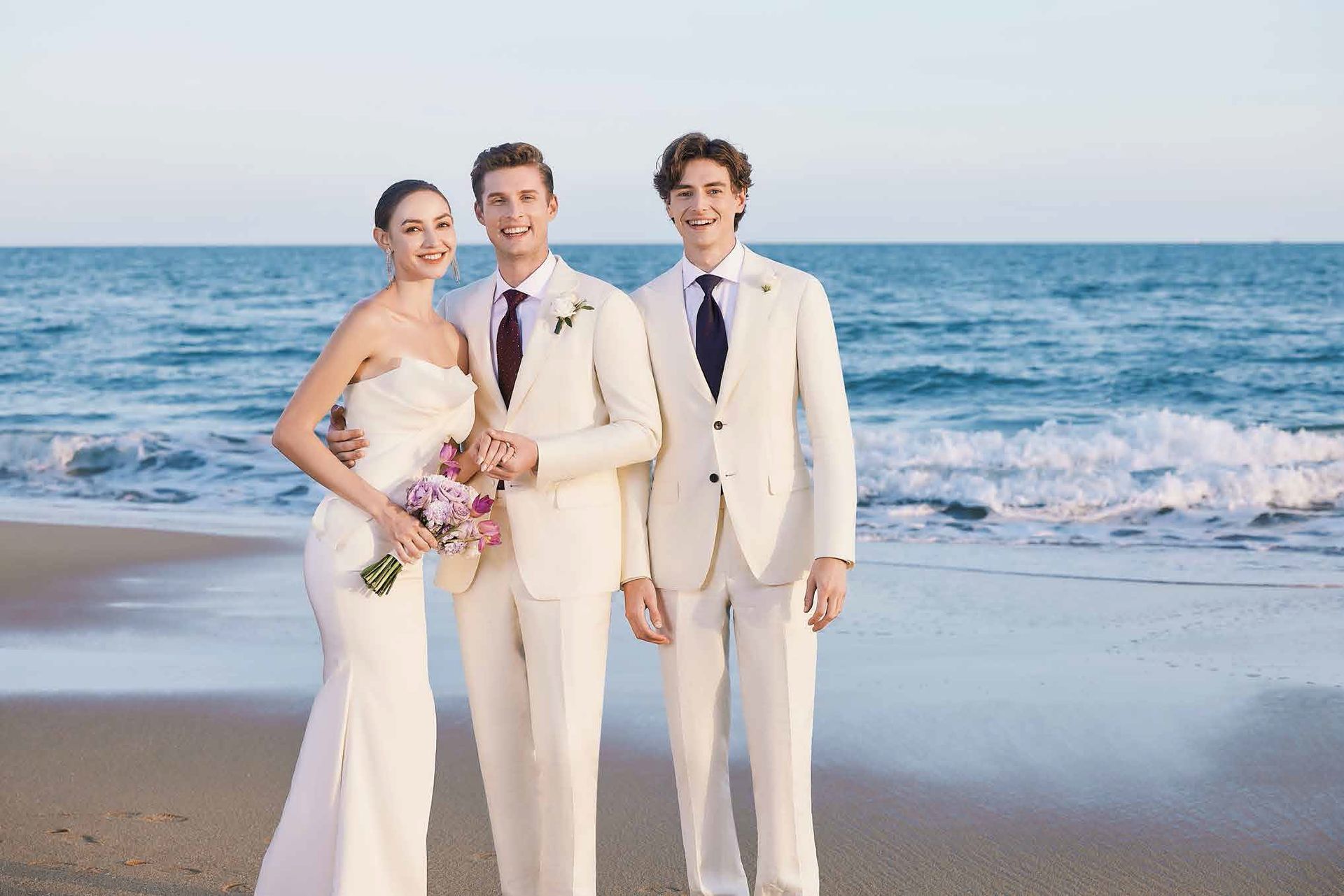 A bride and groom are posing for a picture on the beach with two men in suits.
