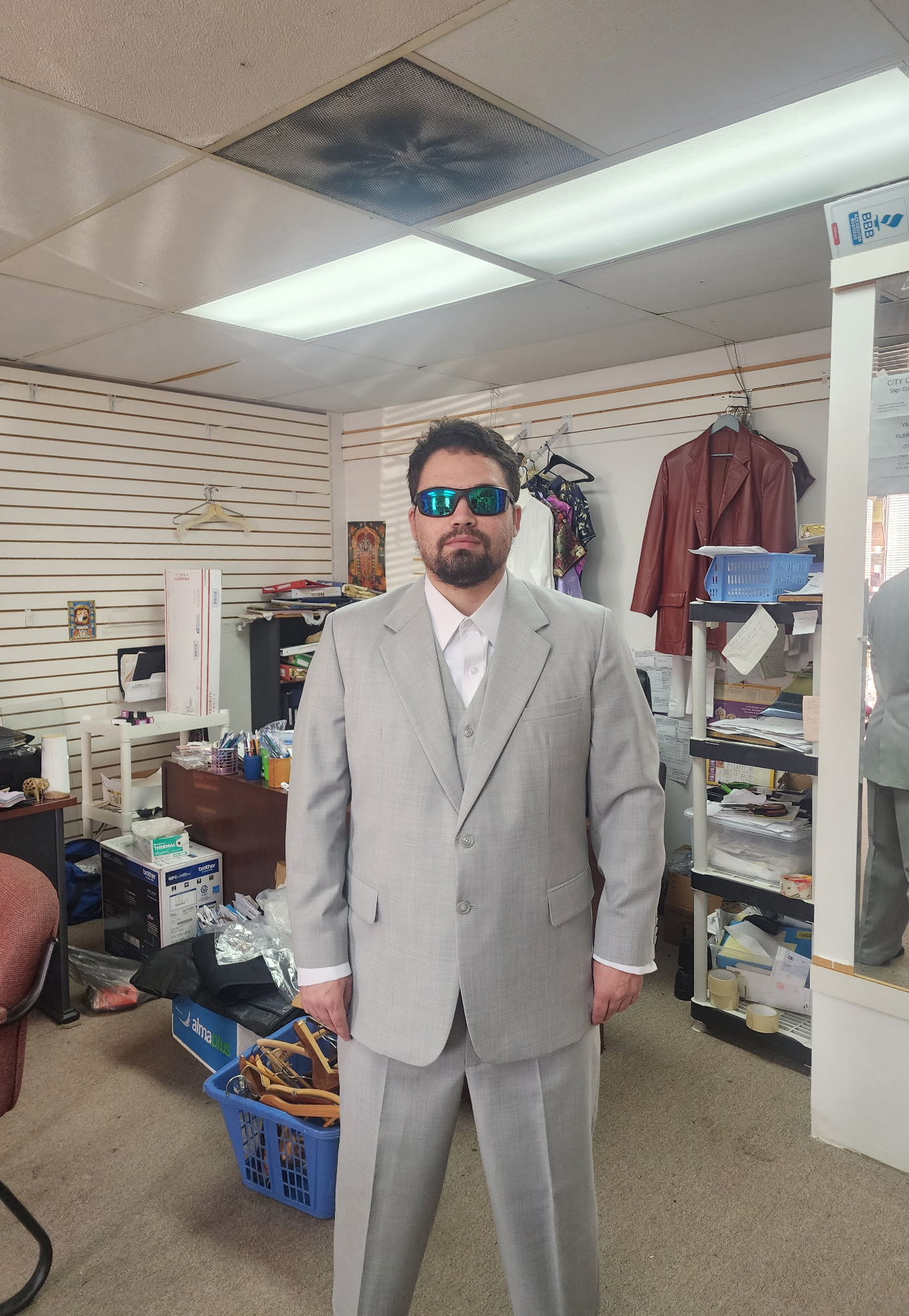 Man in gray suit and sunglasses stands inside a store with clothing racks and shelves.
