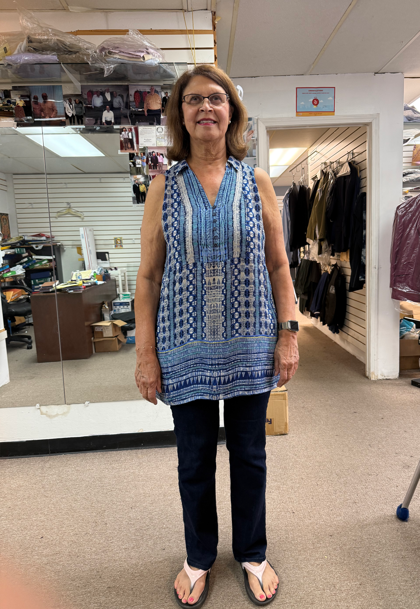 Woman wearing a sleeveless blue patterned top and dark jeans, standing in a store with a mirror.