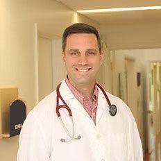 A man in a lab coat with a stethoscope around his neck is standing in a hallway.