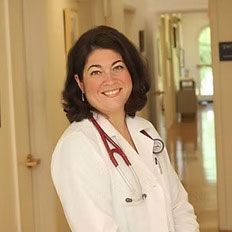 A woman in a lab coat with a stethoscope around her neck is smiling in a hallway.