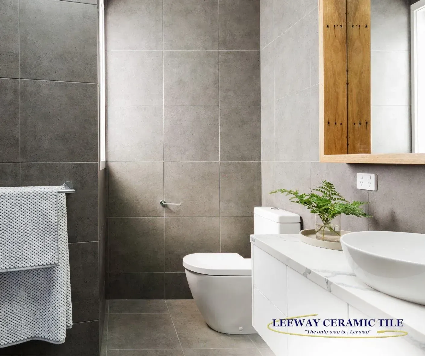 Modern bathroom with gray tiled walls, white toilet and sink, wooden mirror frame.