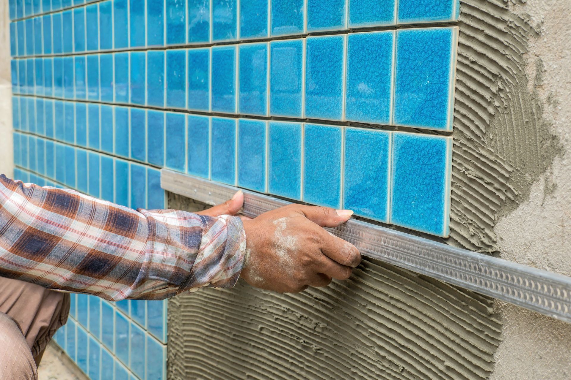 Person tiling blue rectangular tiles onto a wall with mortar, using a level.