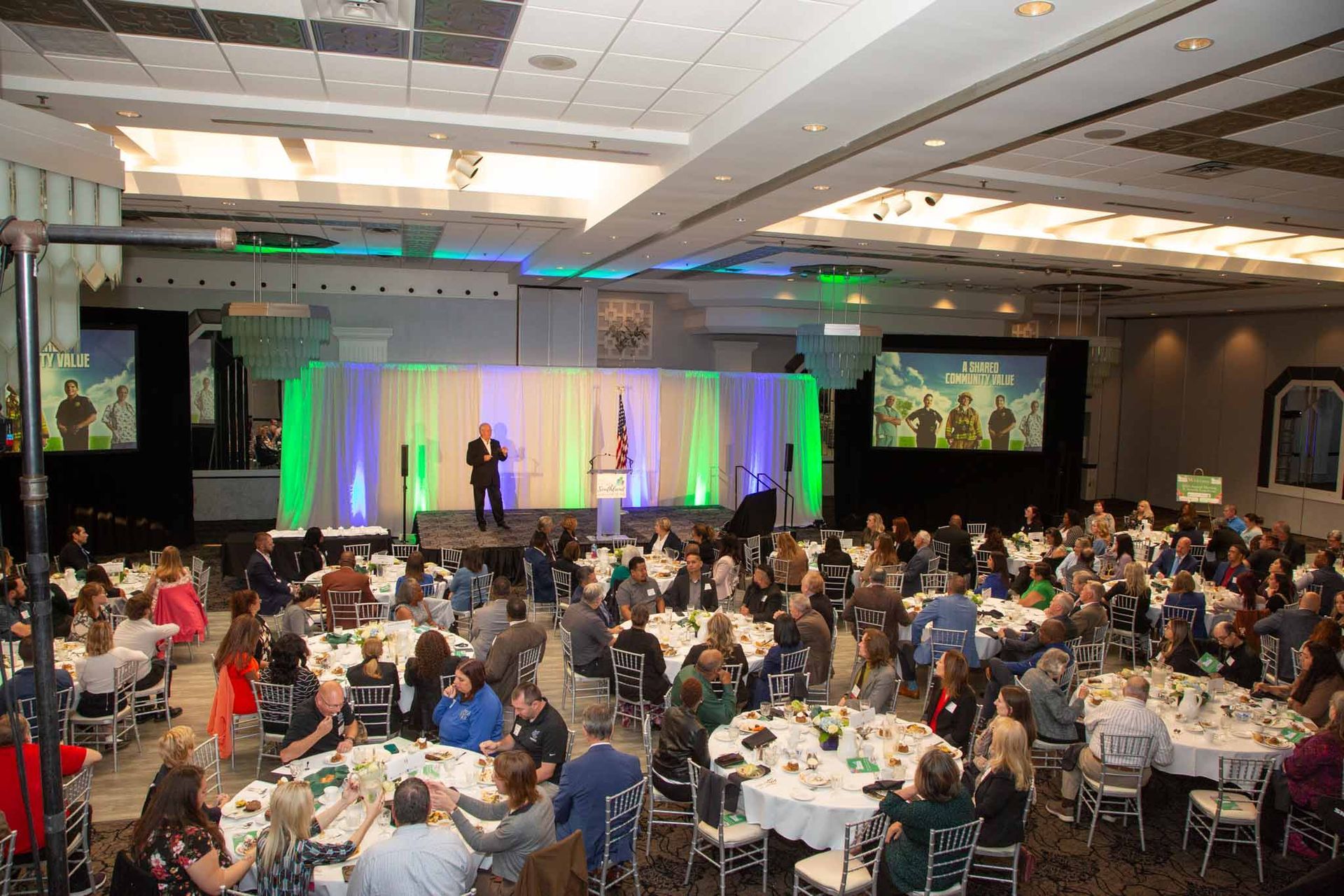 A large banquet hall with people seated at round tables, a speaker on stage with green and blue lighting.