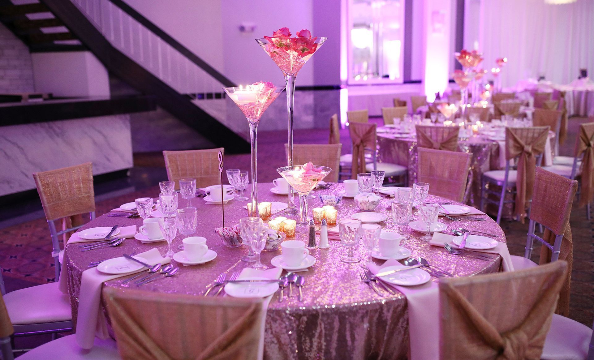 Round tables set for a formal event with pink sequined tablecloths and tall glass centerpieces.