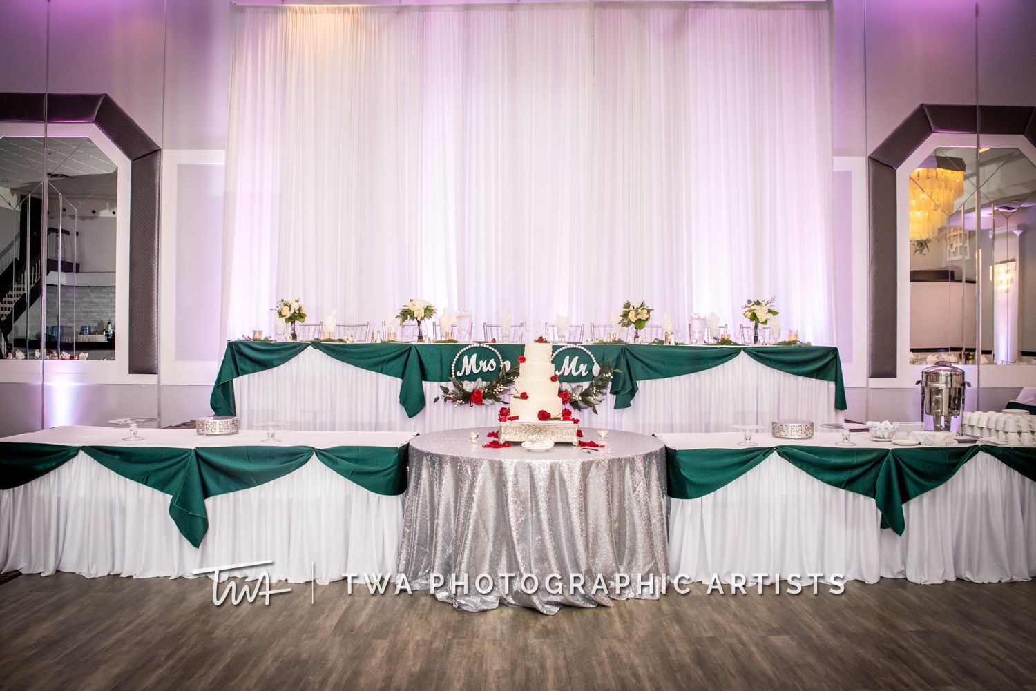 A wedding reception with a cake on a table in front of a white and green backdrop.