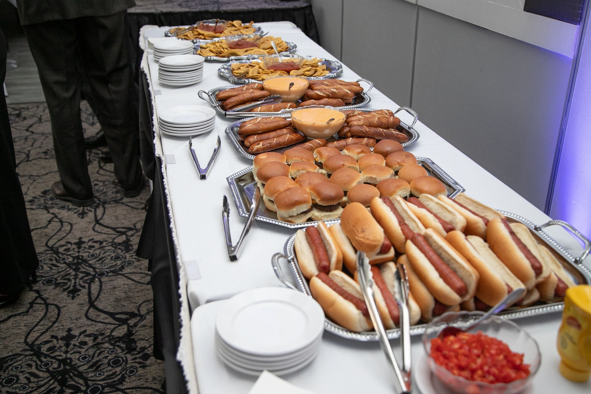 Buffet table with hot dogs, sliders, pretzels, and toppings.