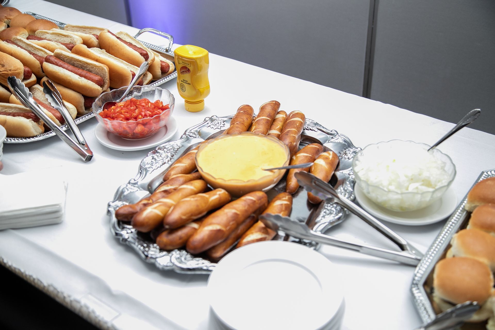 A buffet with hot dogs, pretzels with cheese dip, chopped tomatoes, onions, mustard, and buns.
