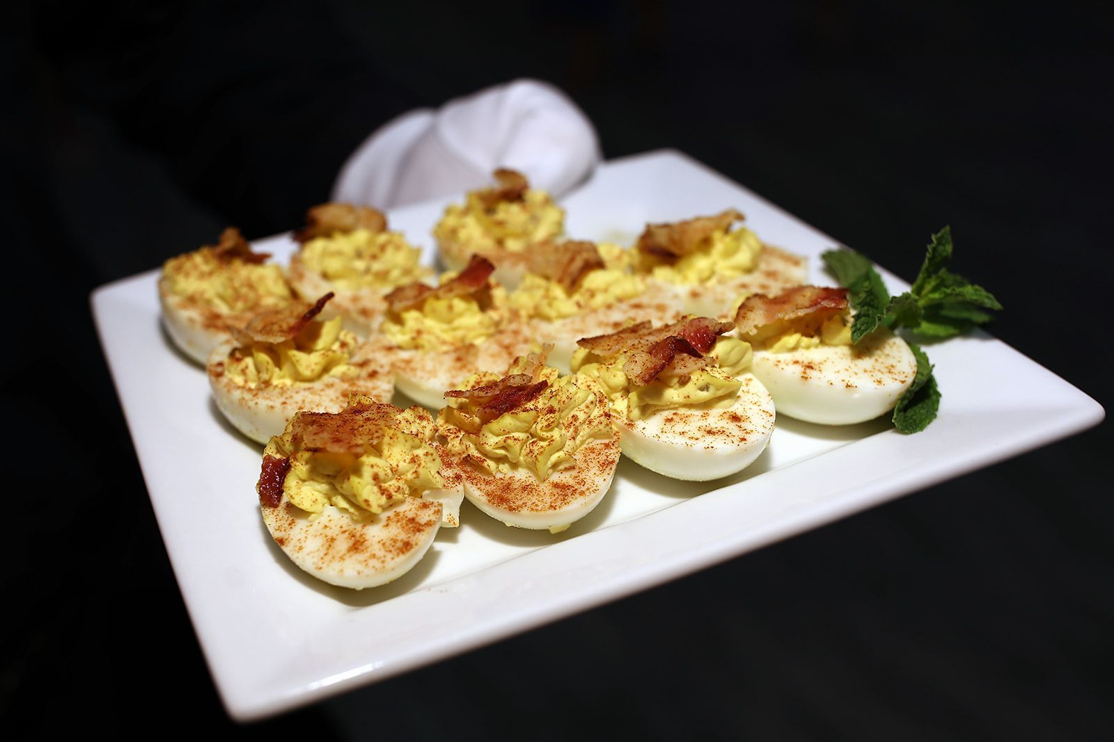 Deviled eggs on a white platter, topped with bacon and paprika, with a sprig of mint.