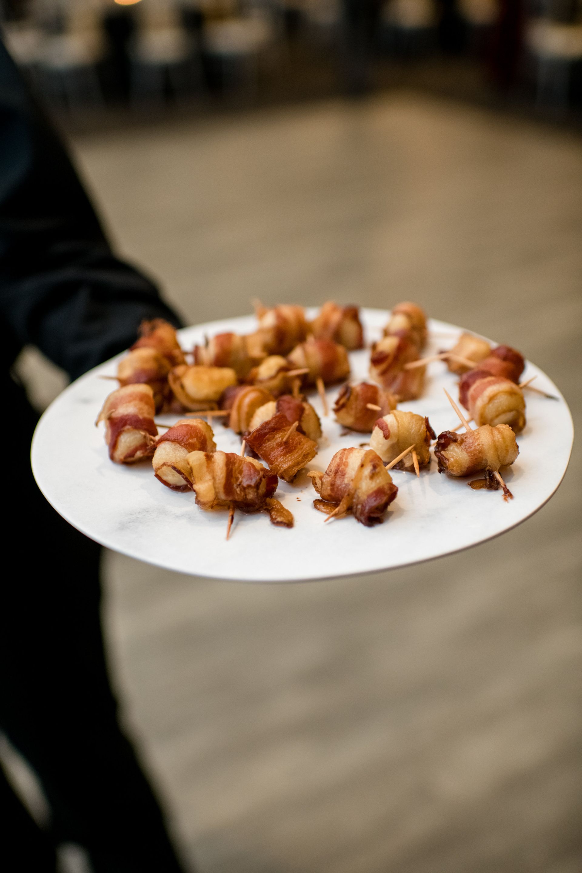 A person holds a white plate of bacon-wrapped appetizers at an event.