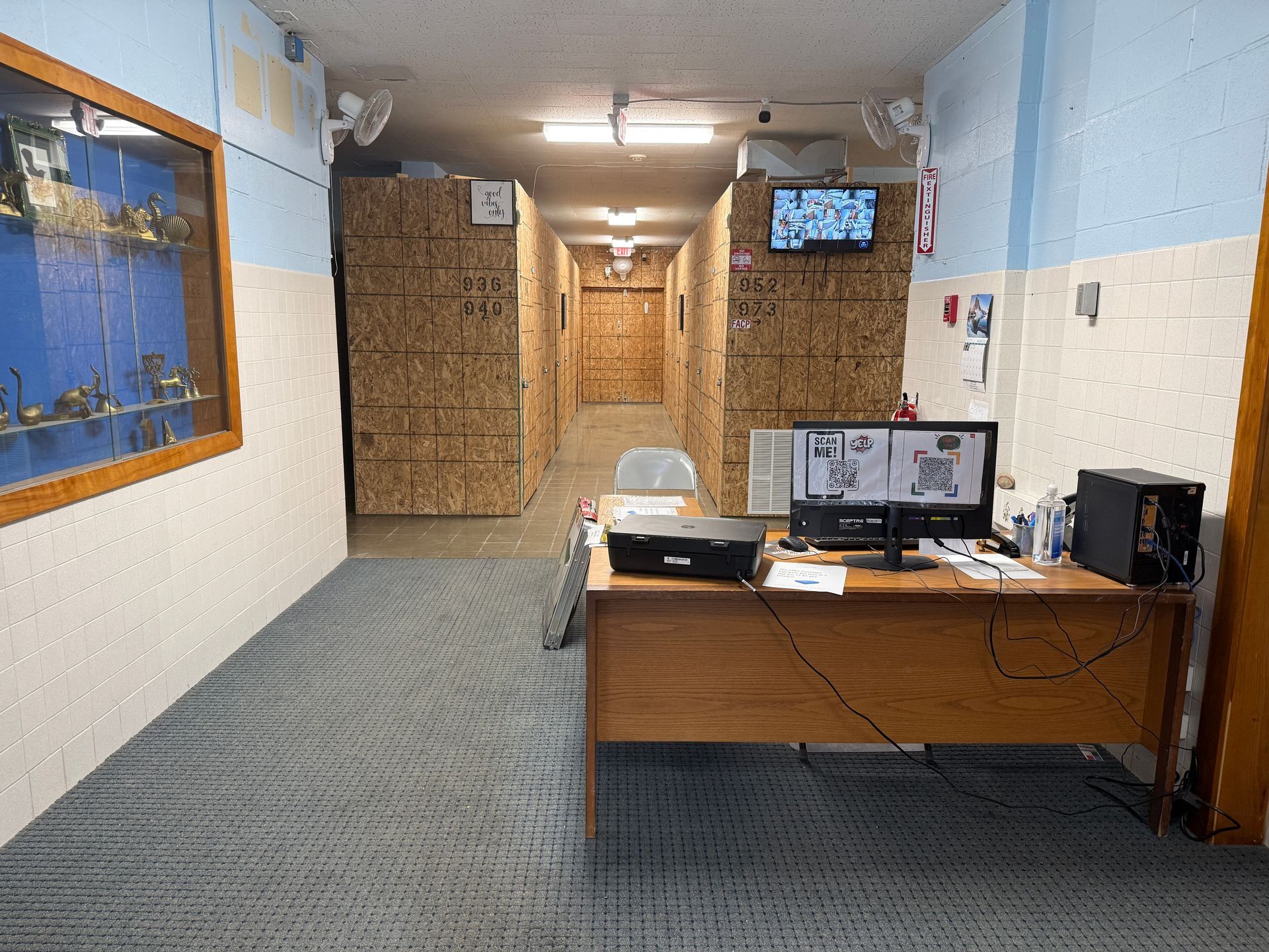 Hallway with desk, computer screens, display case, and security cameras. Corrugated cardboard walls.