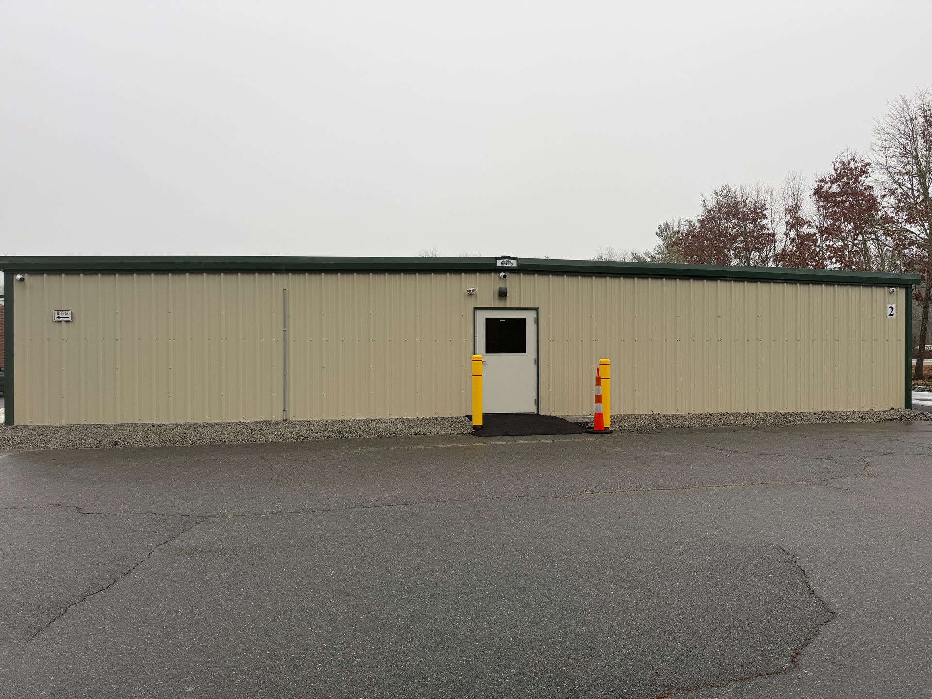 Tan metal building with a door and ramp, two orange bollards, and asphalt parking area.