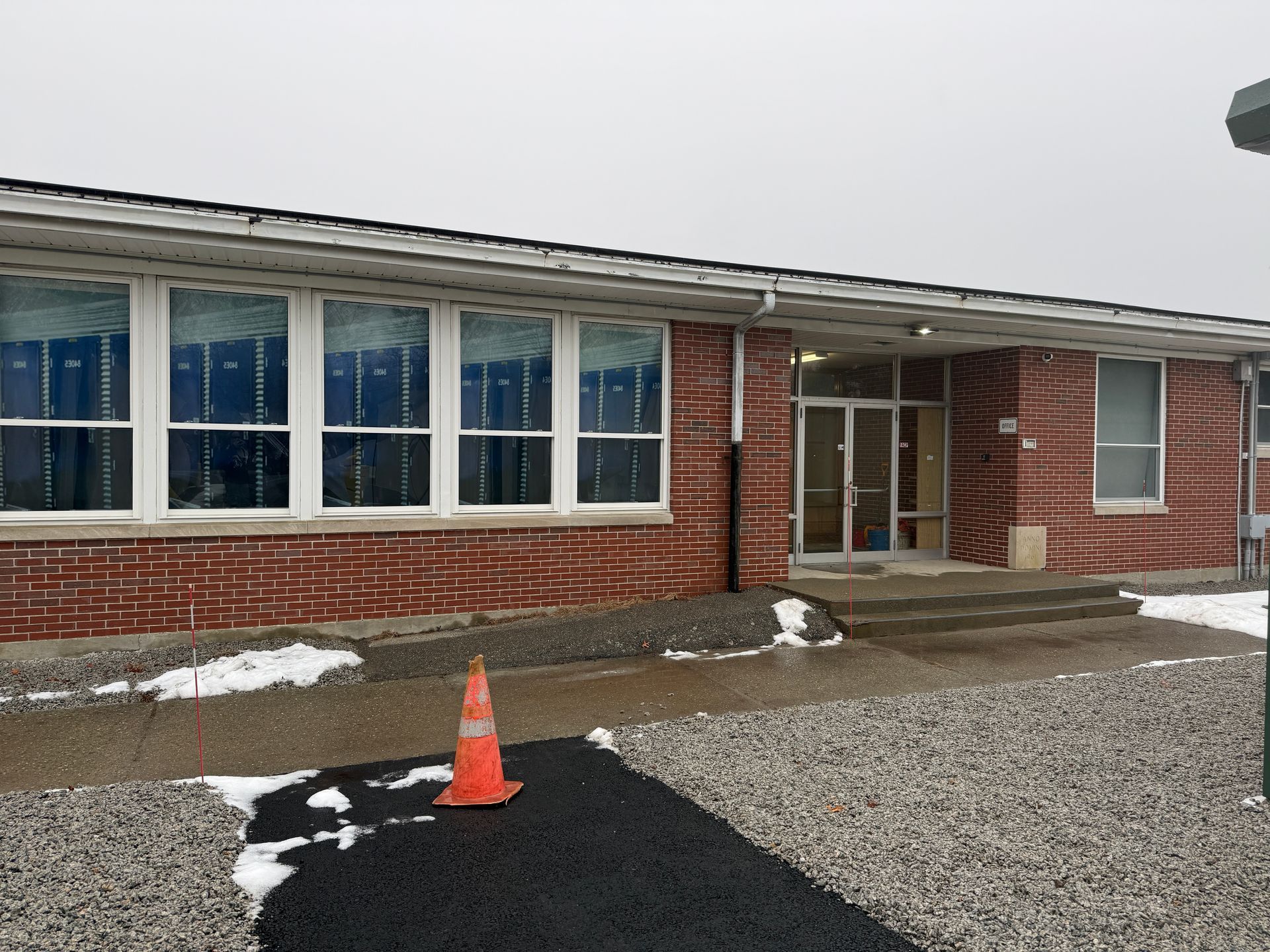 Exterior view of a brick building with windows and a doorway. A traffic cone is on a dark asphalt path next to snow.