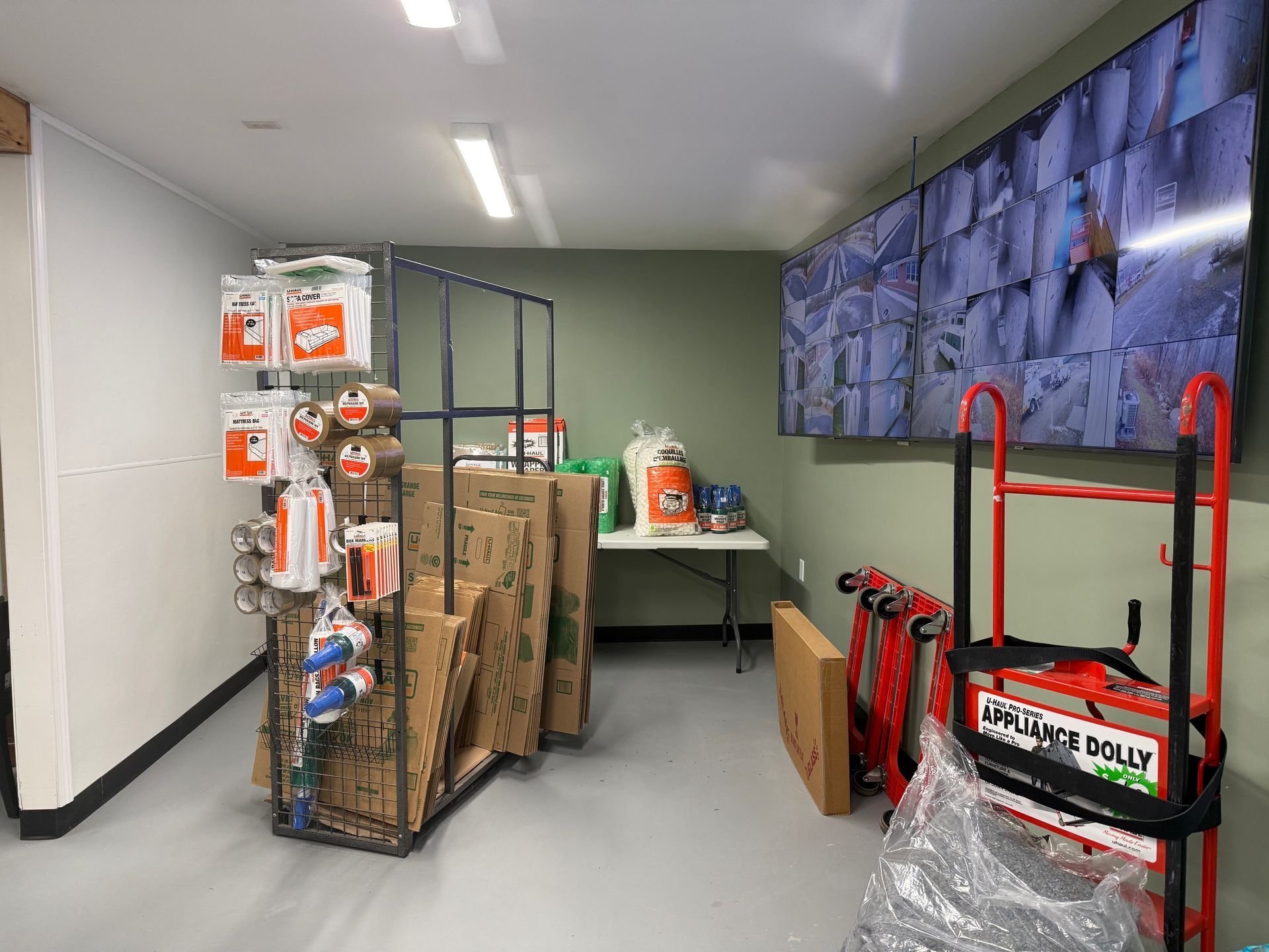 Storage facility interior with packing supplies, a rolling cart, and security monitors.