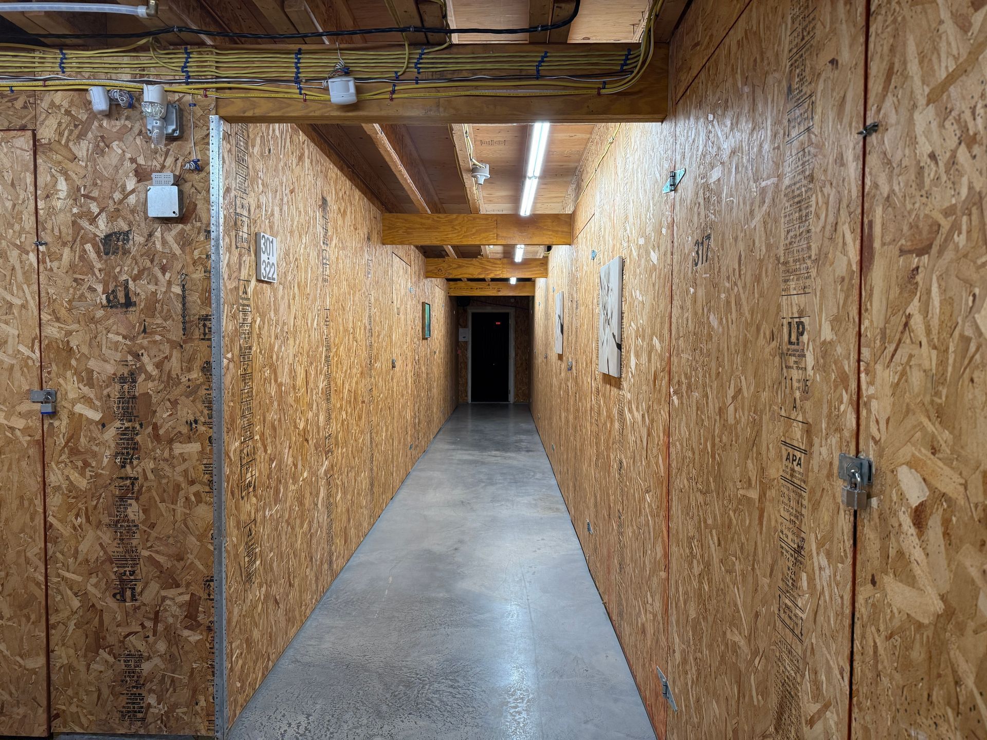 Long, narrow hallway lined with storage units made of wood particle board. Dim lighting.
