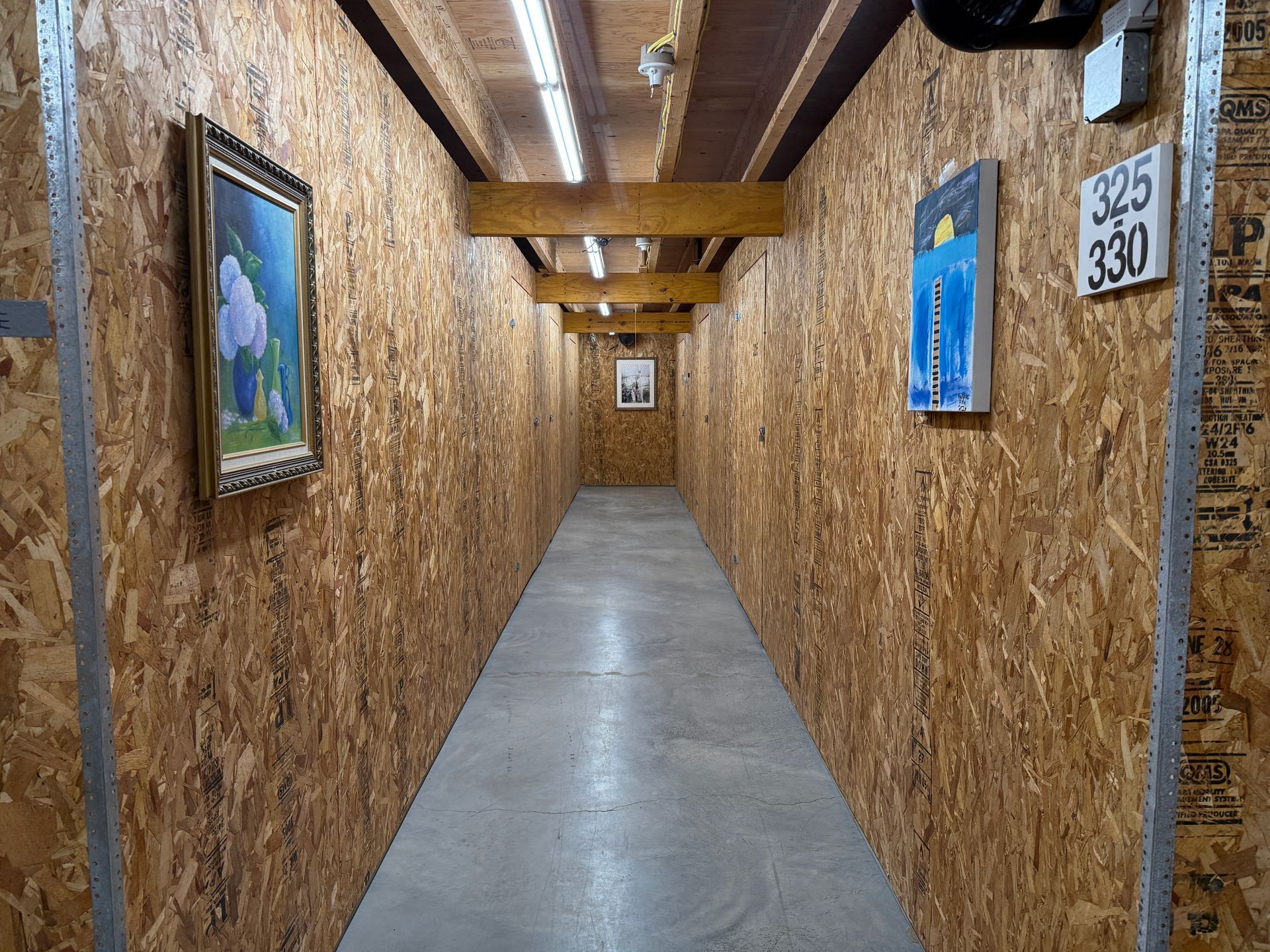 A narrow hallway with art on wood-paneled walls and a concrete floor. Fluorescent lights illuminate the space.