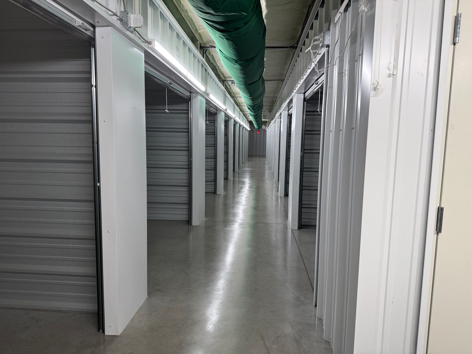 Interior view of a storage unit hallway with open unit doors and fluorescent lighting.