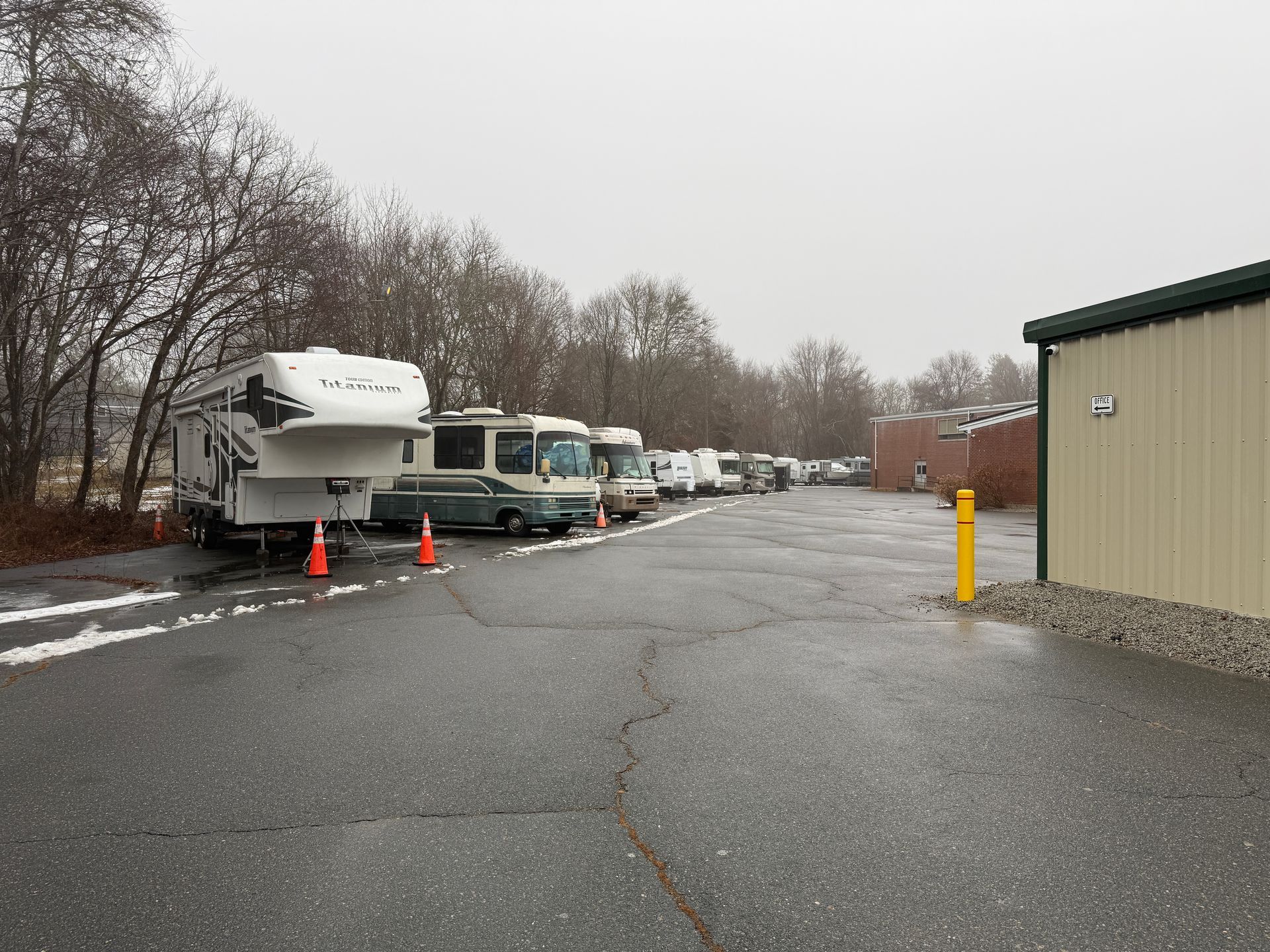 RVs and trailers parked in a lot, gray asphalt, building on the right, trees in the background.