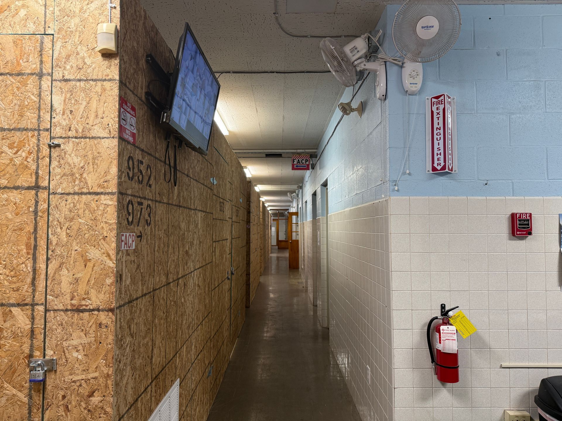 Narrow, long hallway with plywood wall on the left, blue and white tiled wall on the right. A television screen, fan, and fire extinguisher are visible.