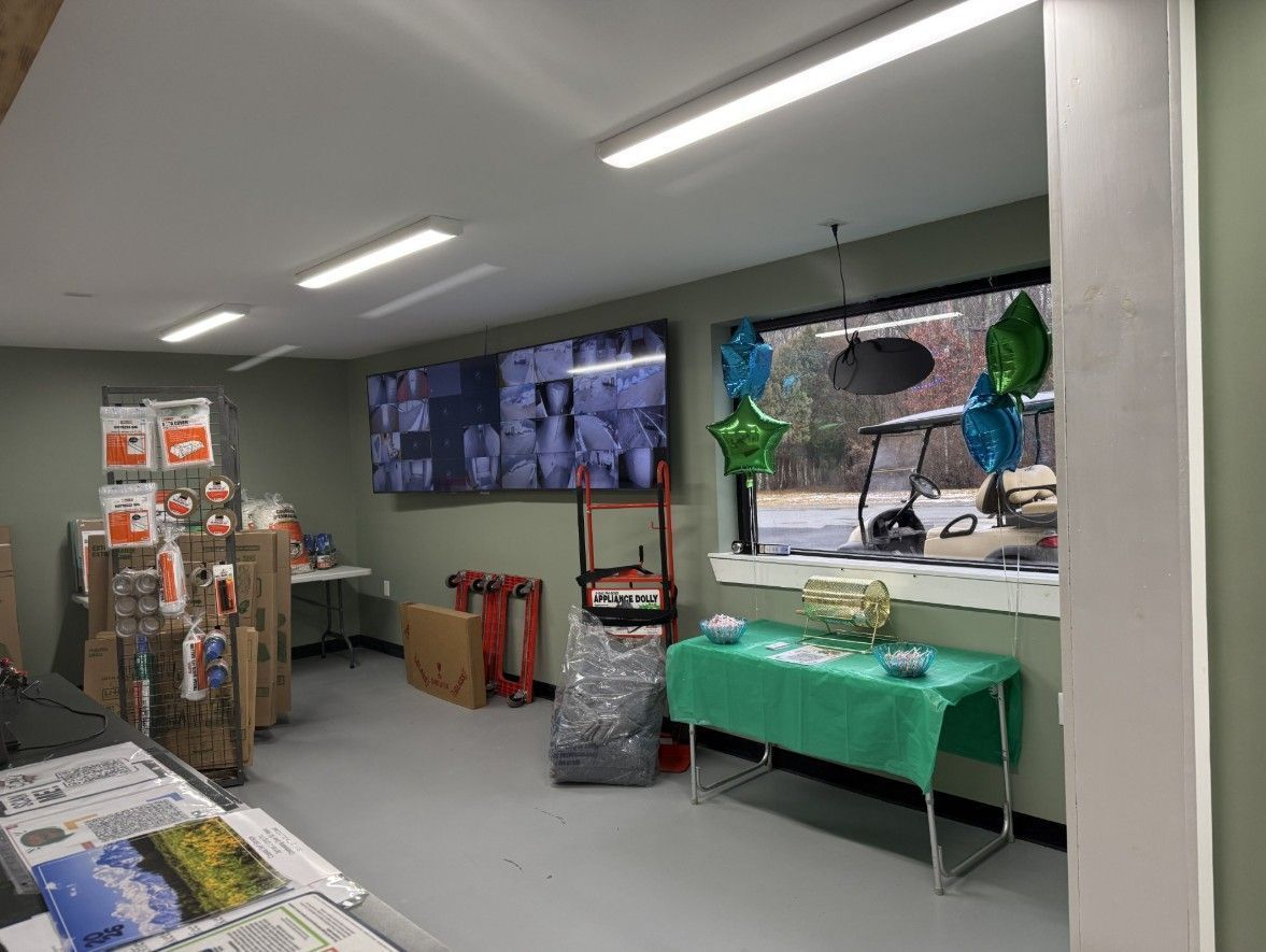 Interior of a store with display shelves, a table, and a monitor showing security footage. A golf cart is seen through a window.