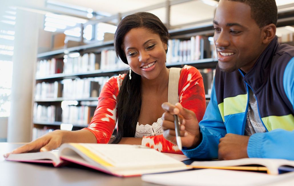 Two people, sitting at a table in a library, looking at a book and smiling.
