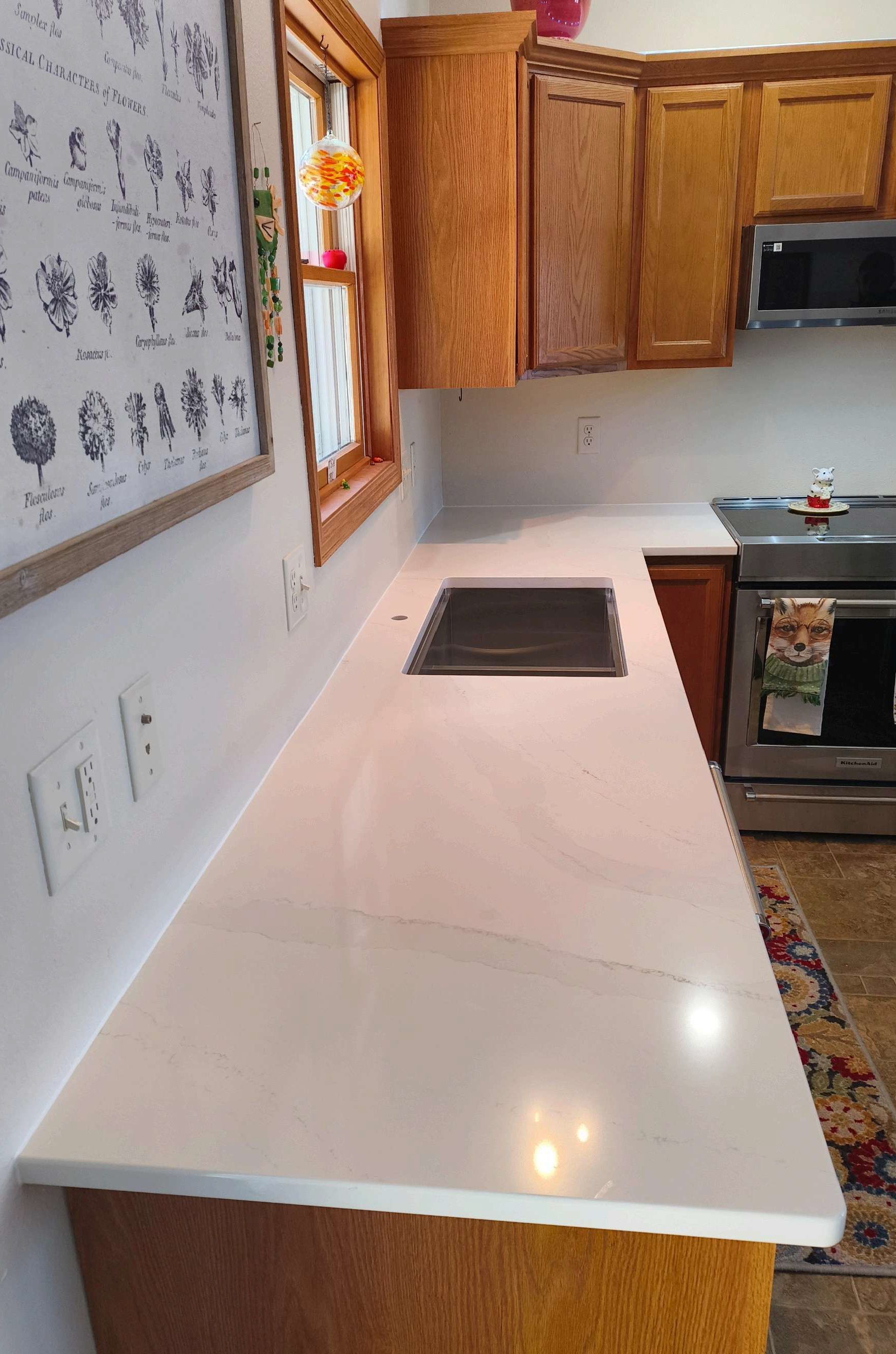 Bright kitchen with a white countertop, stove, wood cabinets, and wall art near a window.