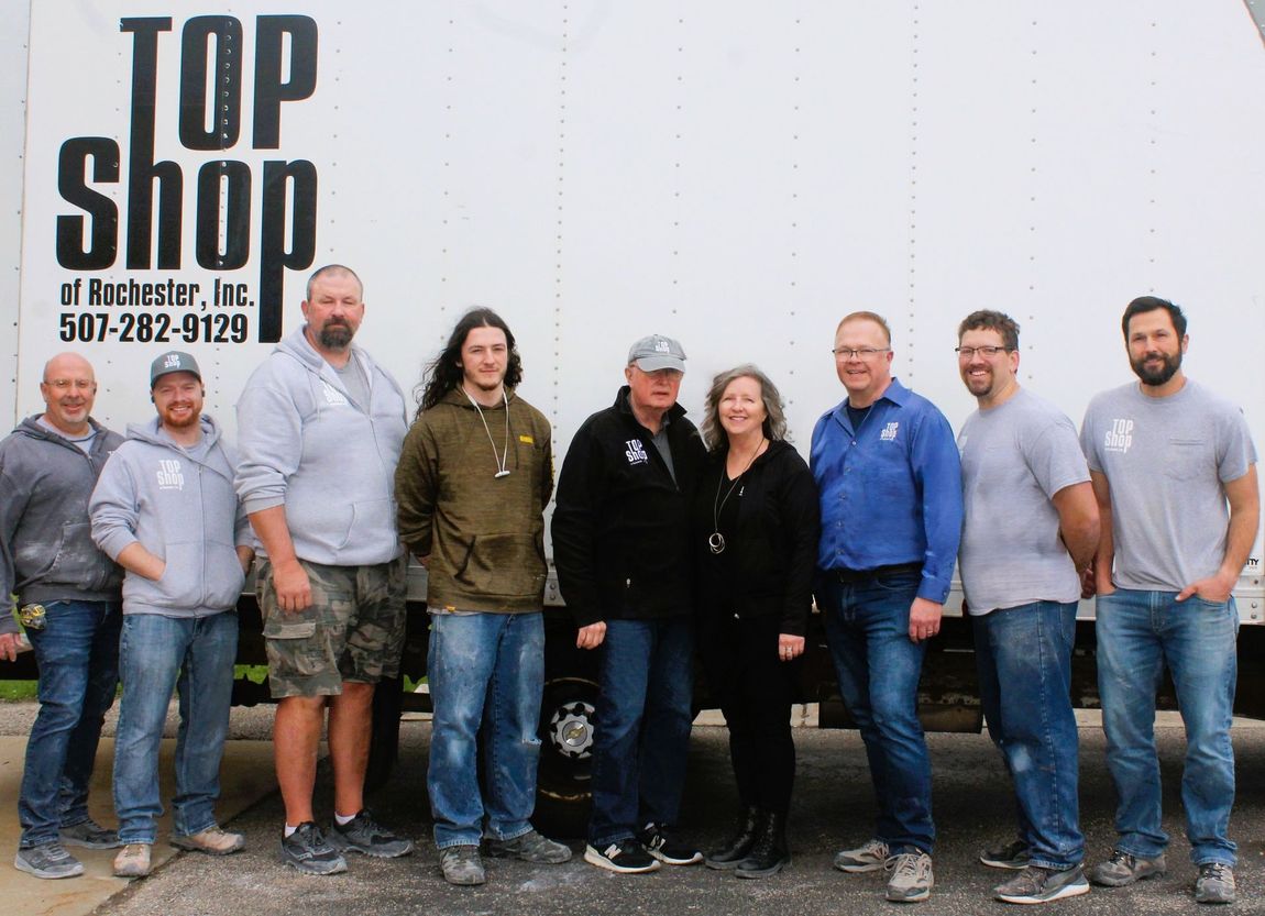 Group of people standing in front of a white truck.  The truck has TOP SHOP and a phone number on it.