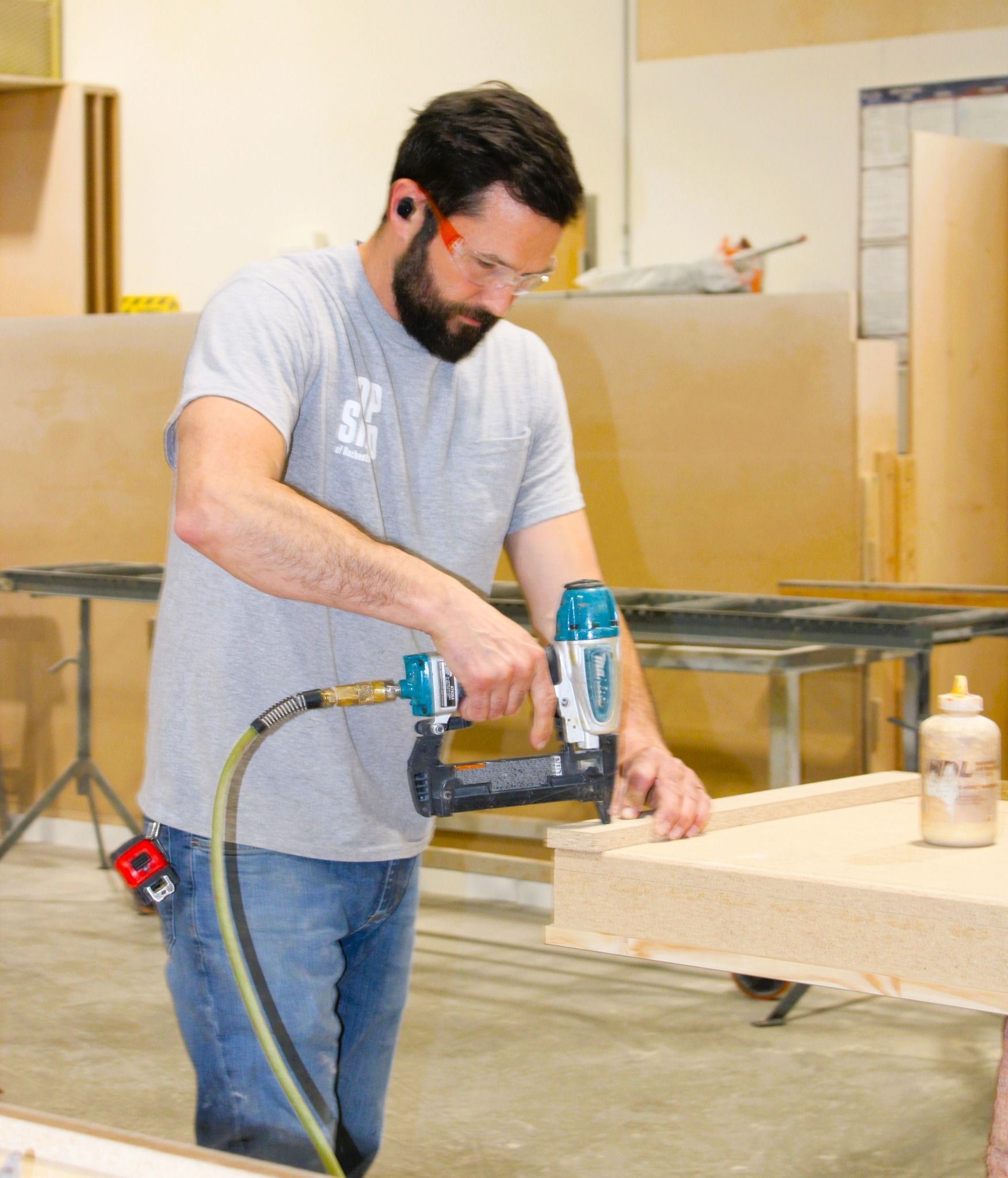 Man using a nail gun on a wood project in a workshop, wearing safety glasses and ear protection.