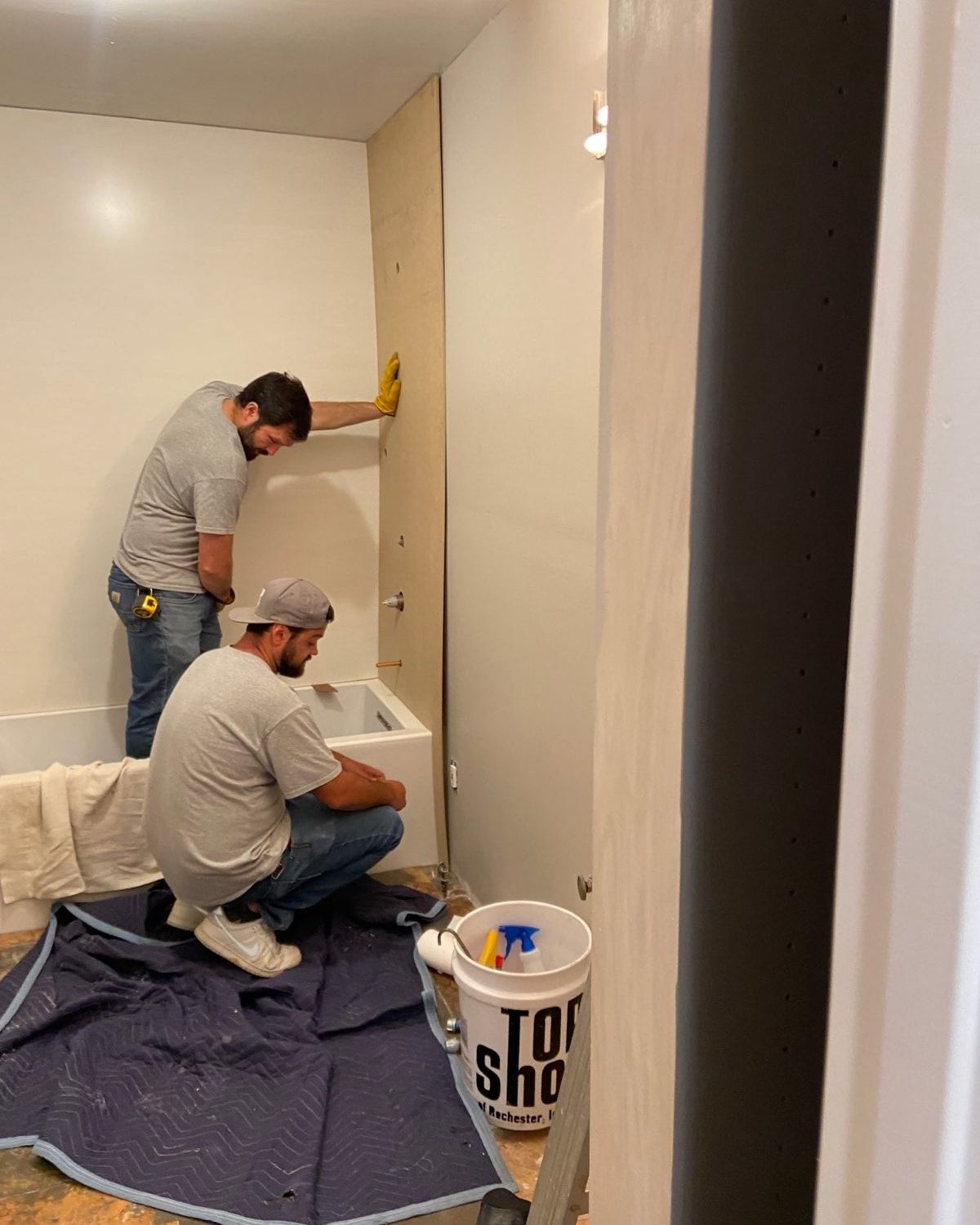 Two workers installing wall panel in a bathroom. One leans, the other kneels near a bathtub. A bucket sits nearby.