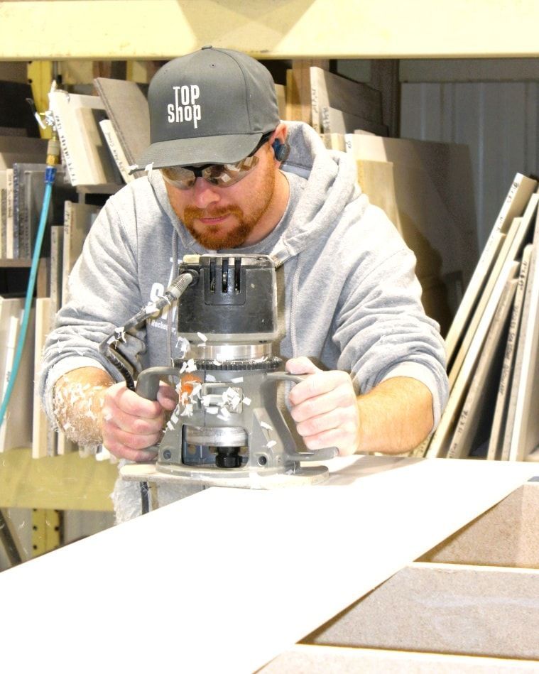 Man in gray hoodie and hat uses a router on a countertop in a workshop.