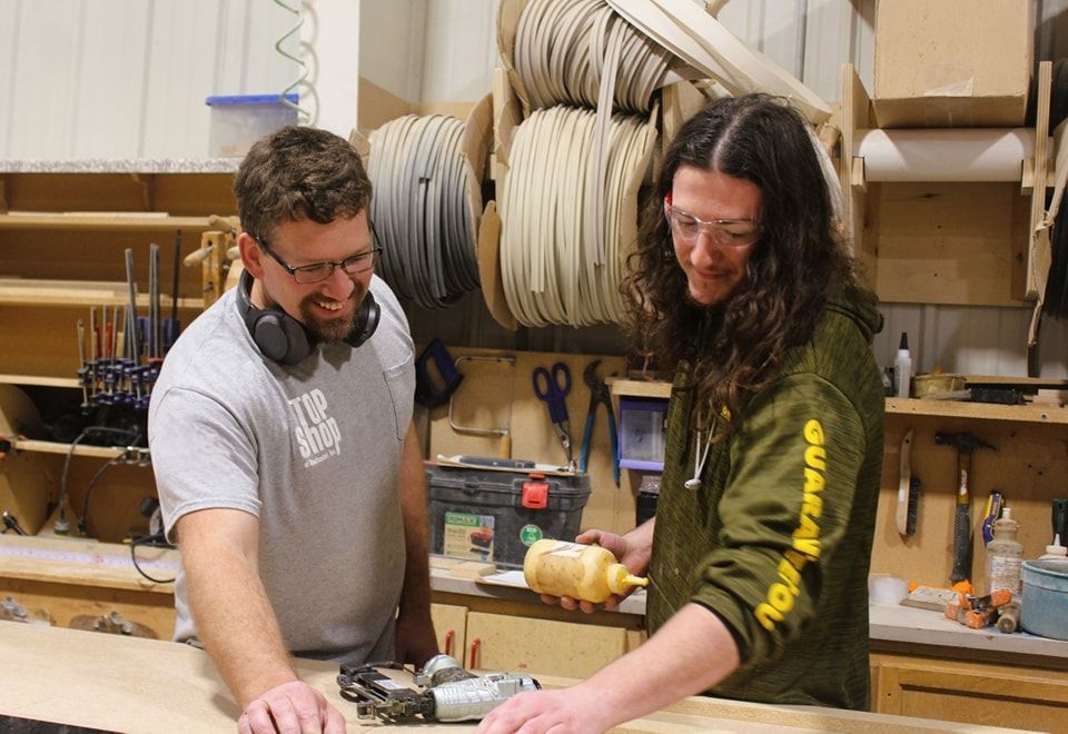 Two people in a workshop, working with wood. One dispenses glue, the other smiles. Tools and materials are in the background.