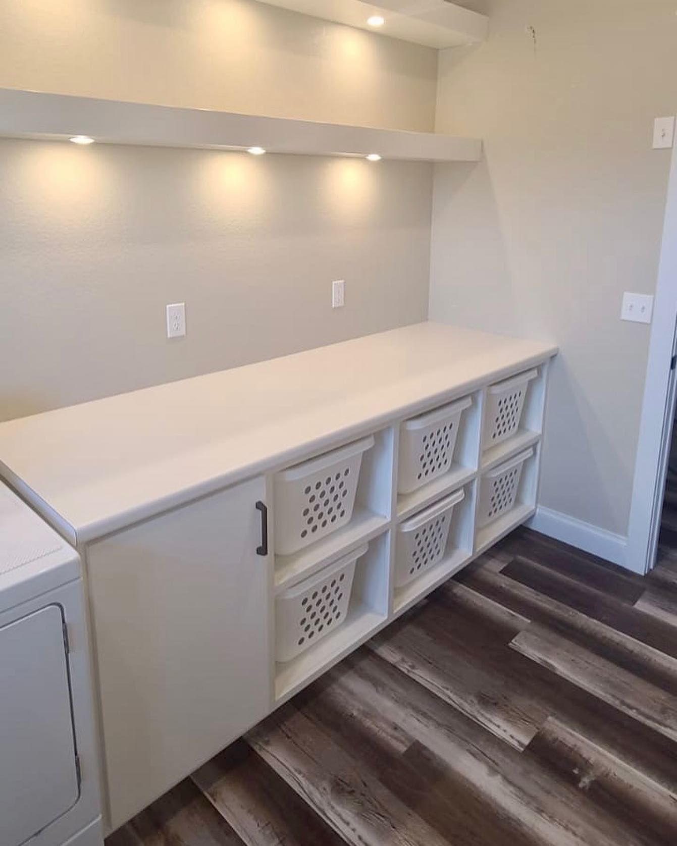 Laundry room with white countertop and built-in baskets, lit by overhead lights. Wooden floor.