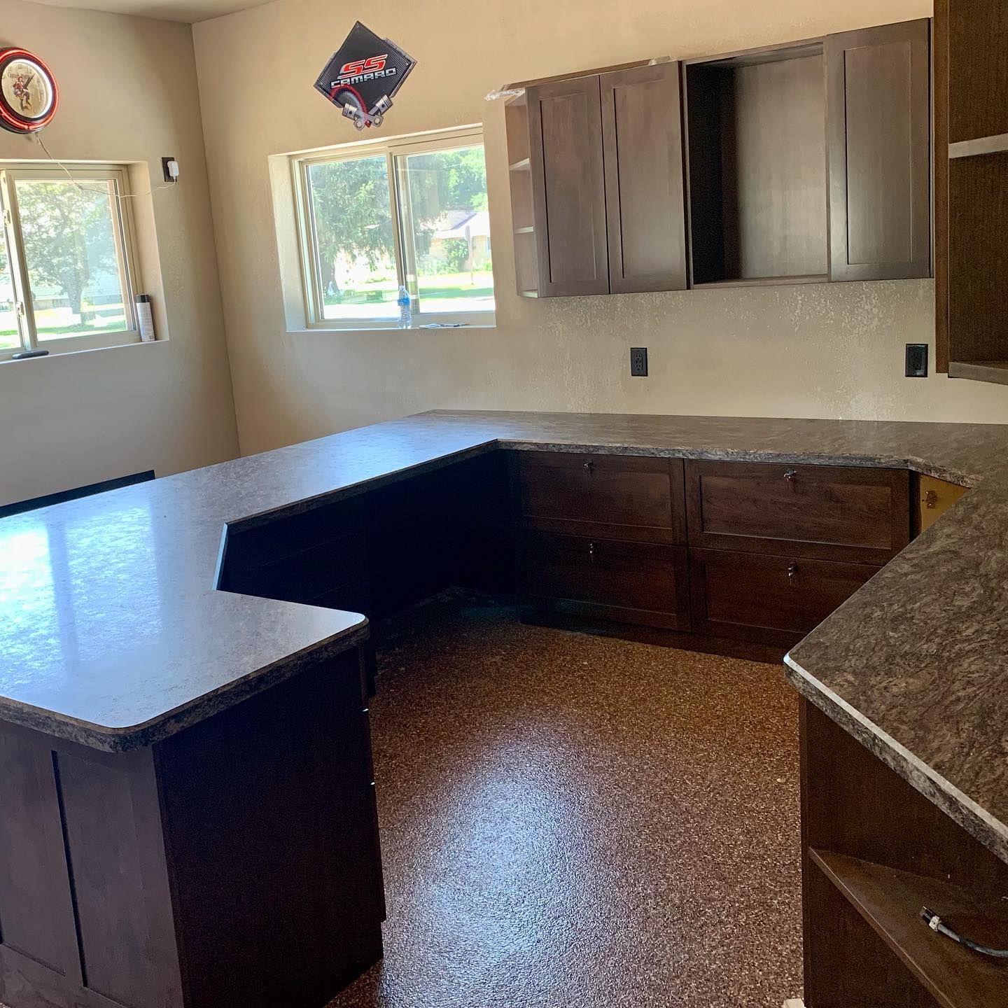 U-shaped kitchen with brown cabinets, gray countertops, and speckled flooring. Two windows and a banner are visible.