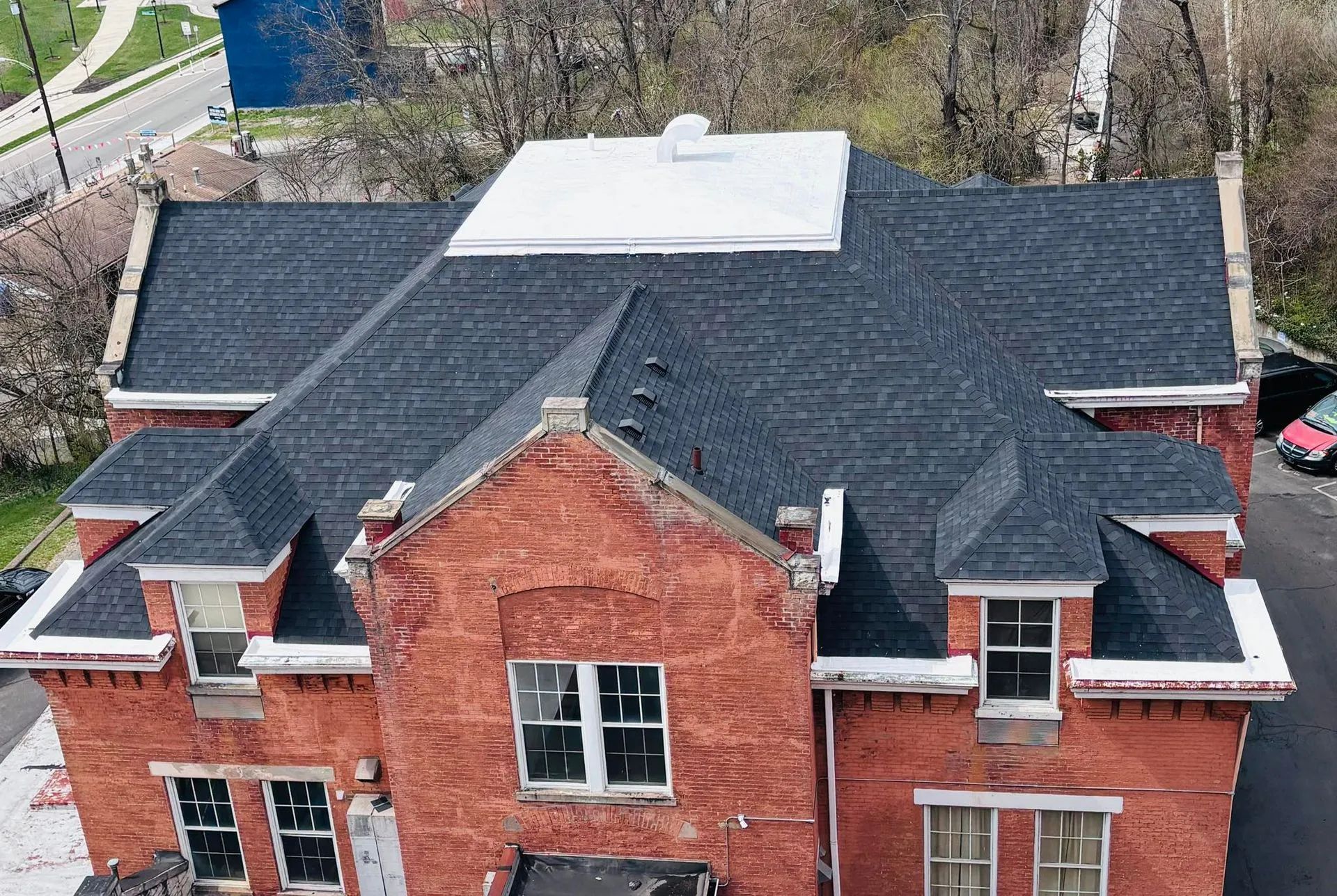 Aerial view of a red brick house with a dark shingled roof and white roof section, surrounded by trees.