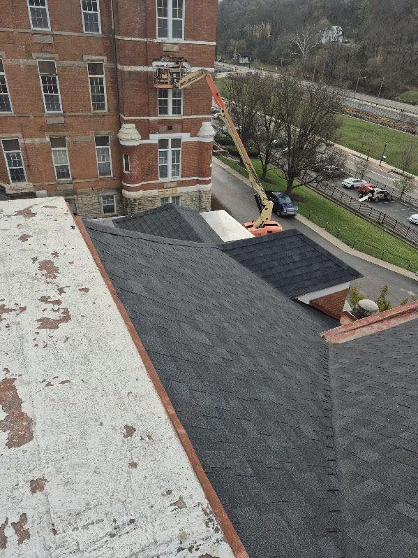 Rooftop with new dark shingles beside an older flat roof and brick building; work truck and trees below