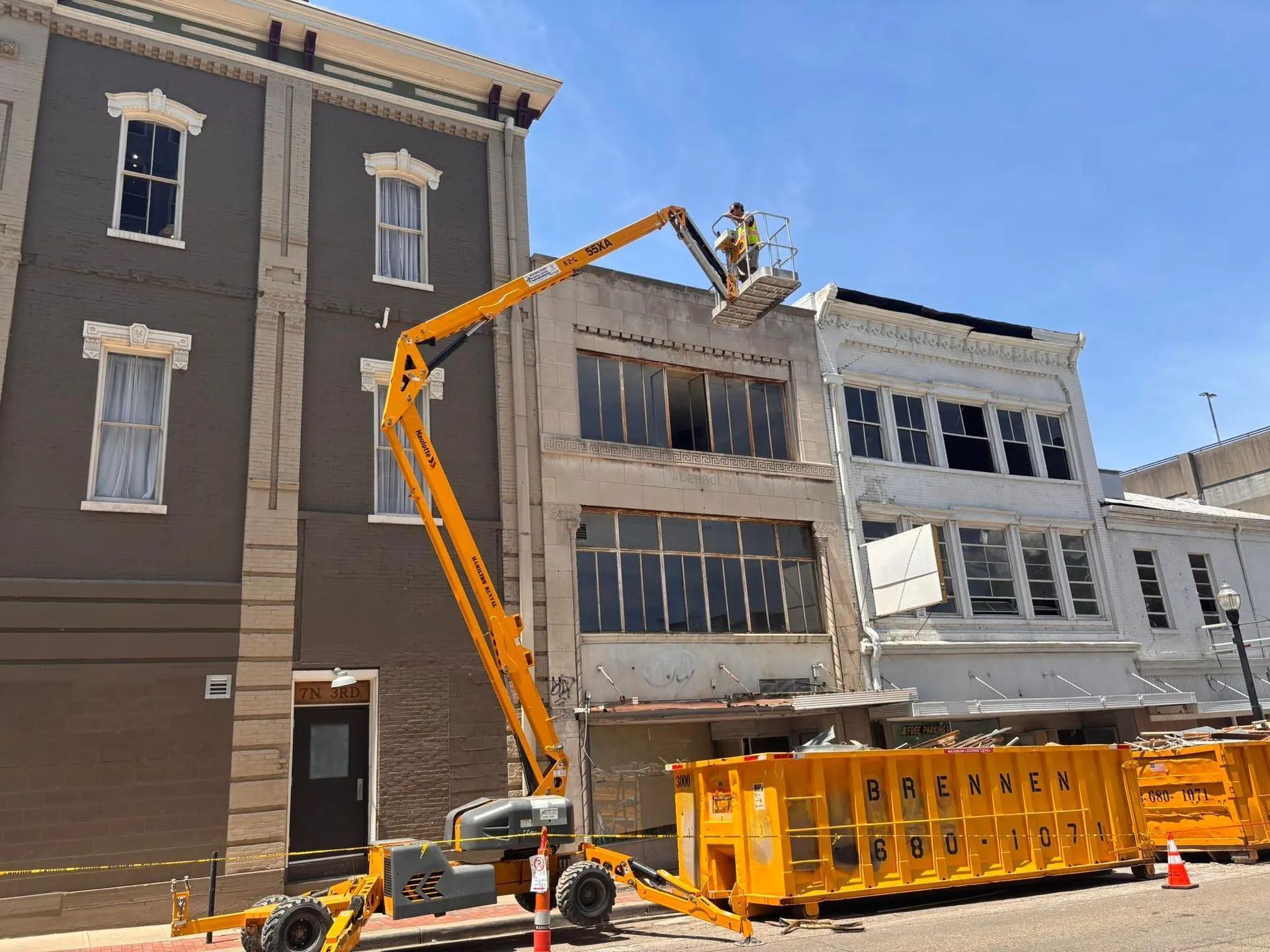 Construction crane demolishing a downtown building beside neighboring facades and a yellow dumpster