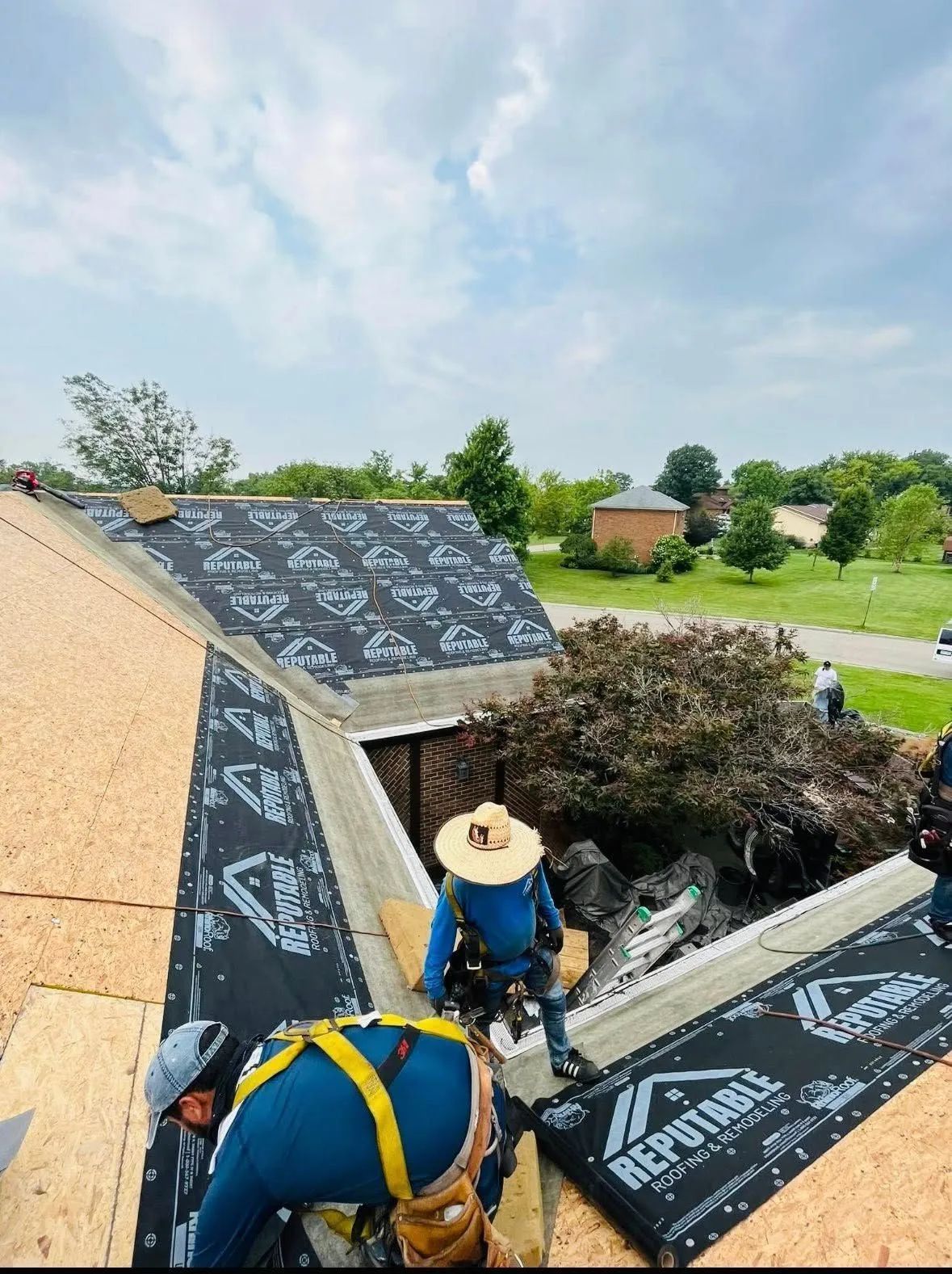 Two roofers working on a roof opening, installing black shingles on a house in a green rural area