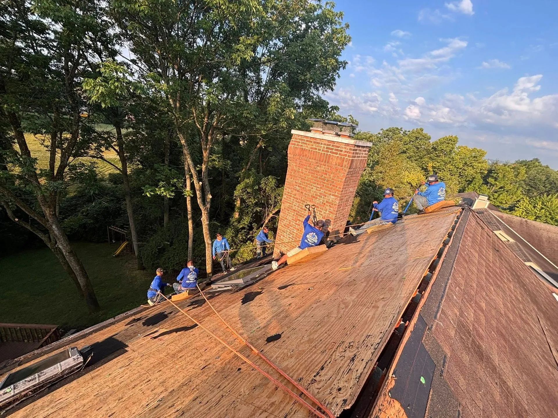 Roofers in blue shirts work on a steep shingled roof beside a brick chimney and trees.