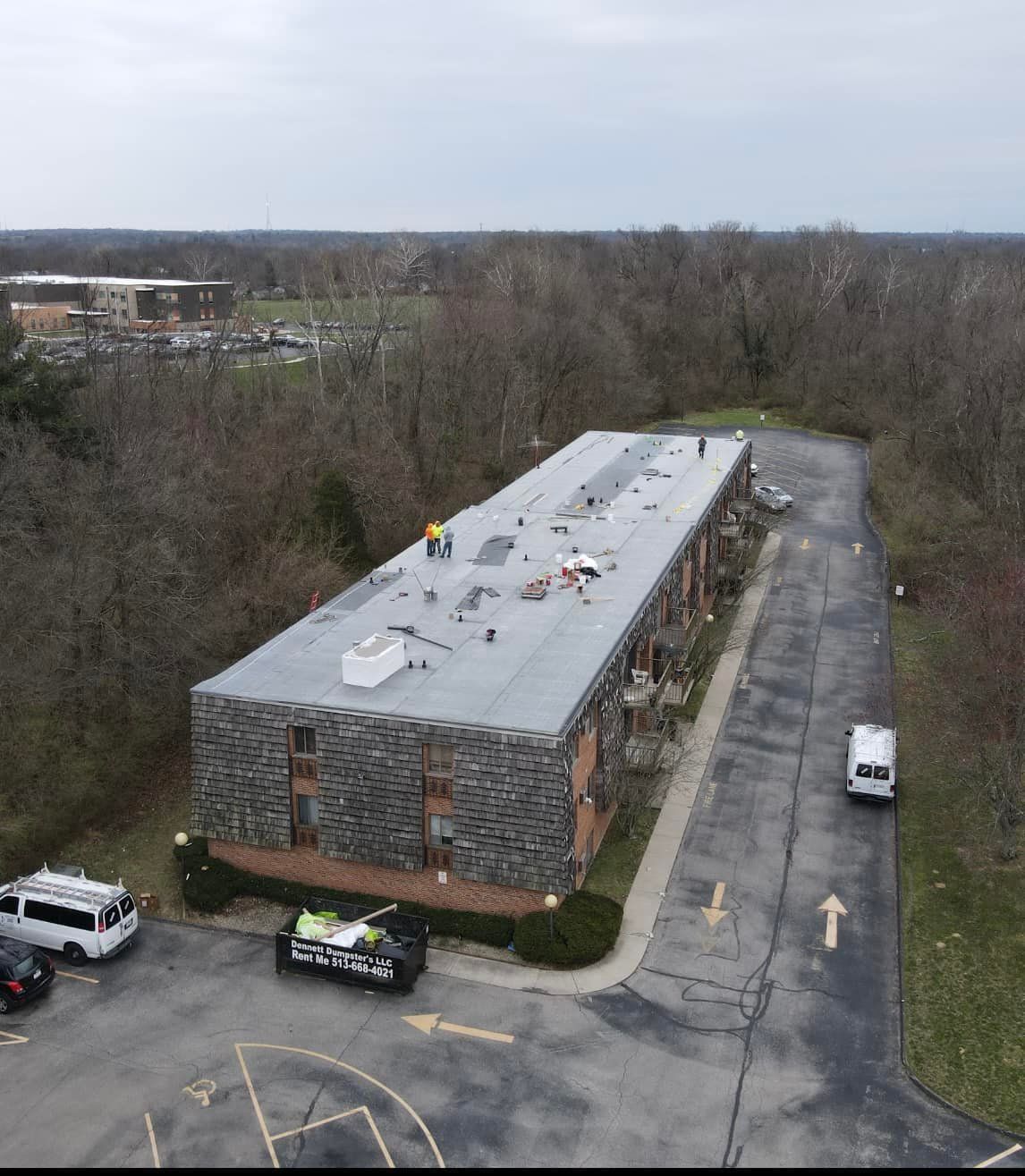 Aerial view of a long flat-roofed building in a parking lot beside trees, with several vehicles nearby.