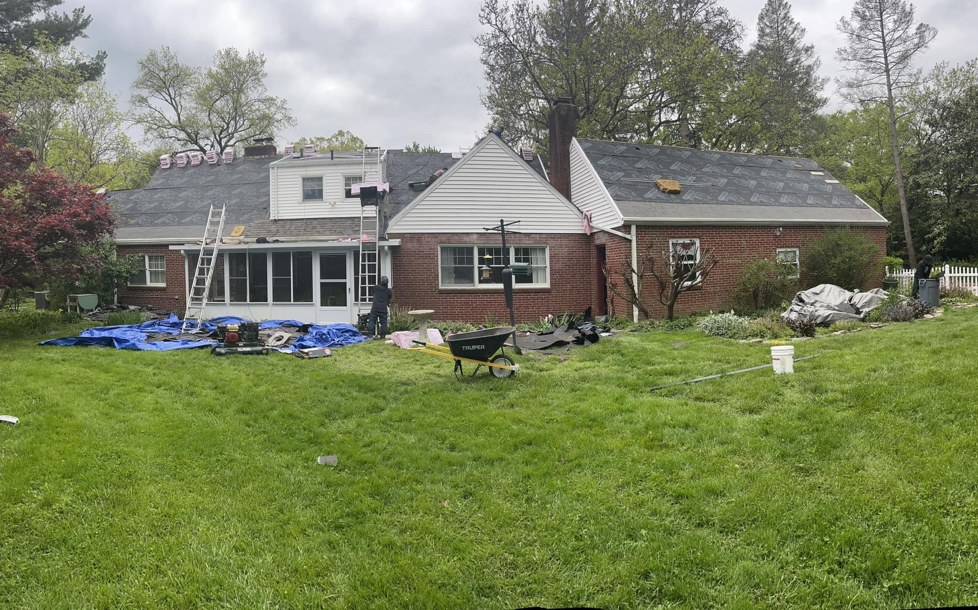 House with damaged roof and yard debris, surrounded by trees and overgrown grass