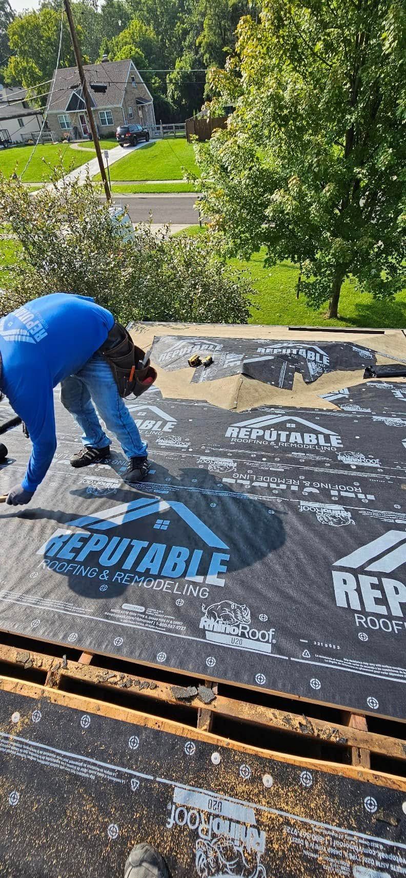 Worker in blue shirt installing roofing felt on a rooftop with trees and houses in the background