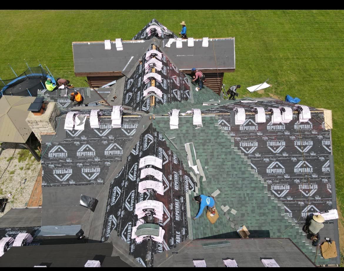 Roof workers installing black shingles on a steep house roof, with tools and materials spread across the surface.