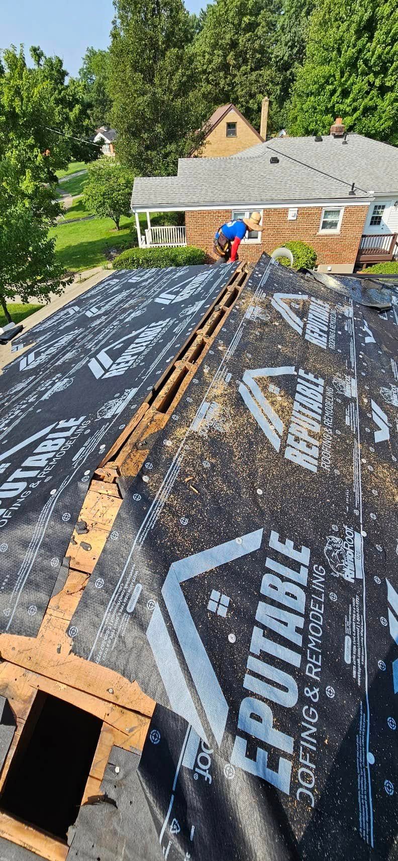 Roofer working on a house roof covered with black underlayment and wooden planks, surrounded by trees