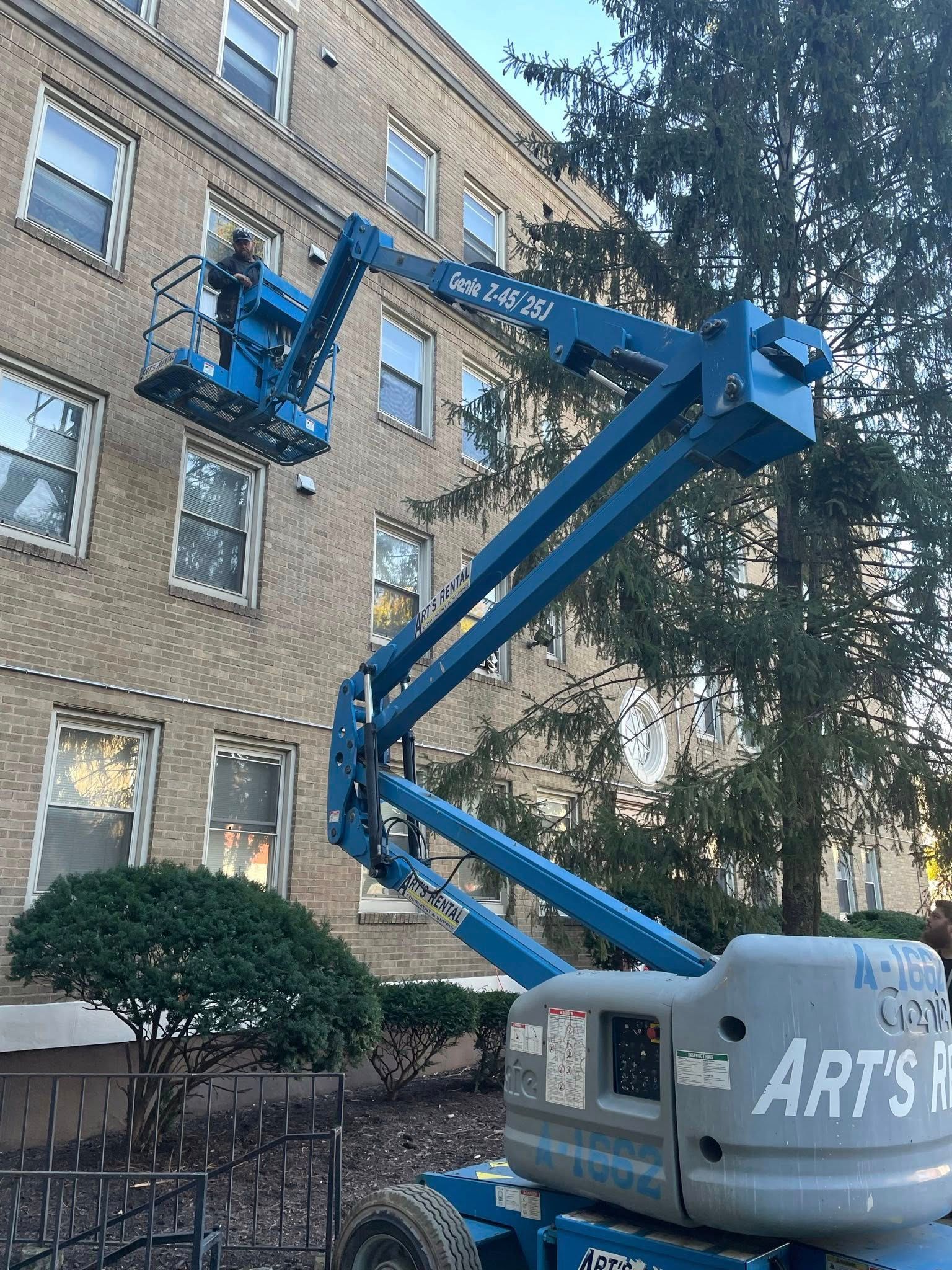 Blue articulated lift raised beside a brick building, reaching toward upper windows and trees