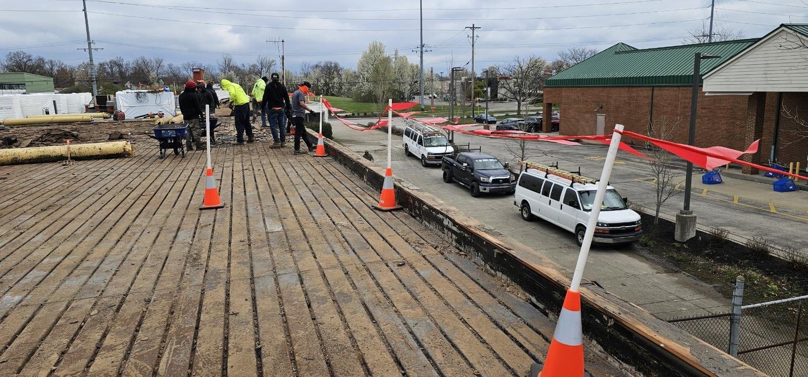 Workers inspect a wooden deck beside parked cars, with orange cones and caution tape under a cloudy sky.